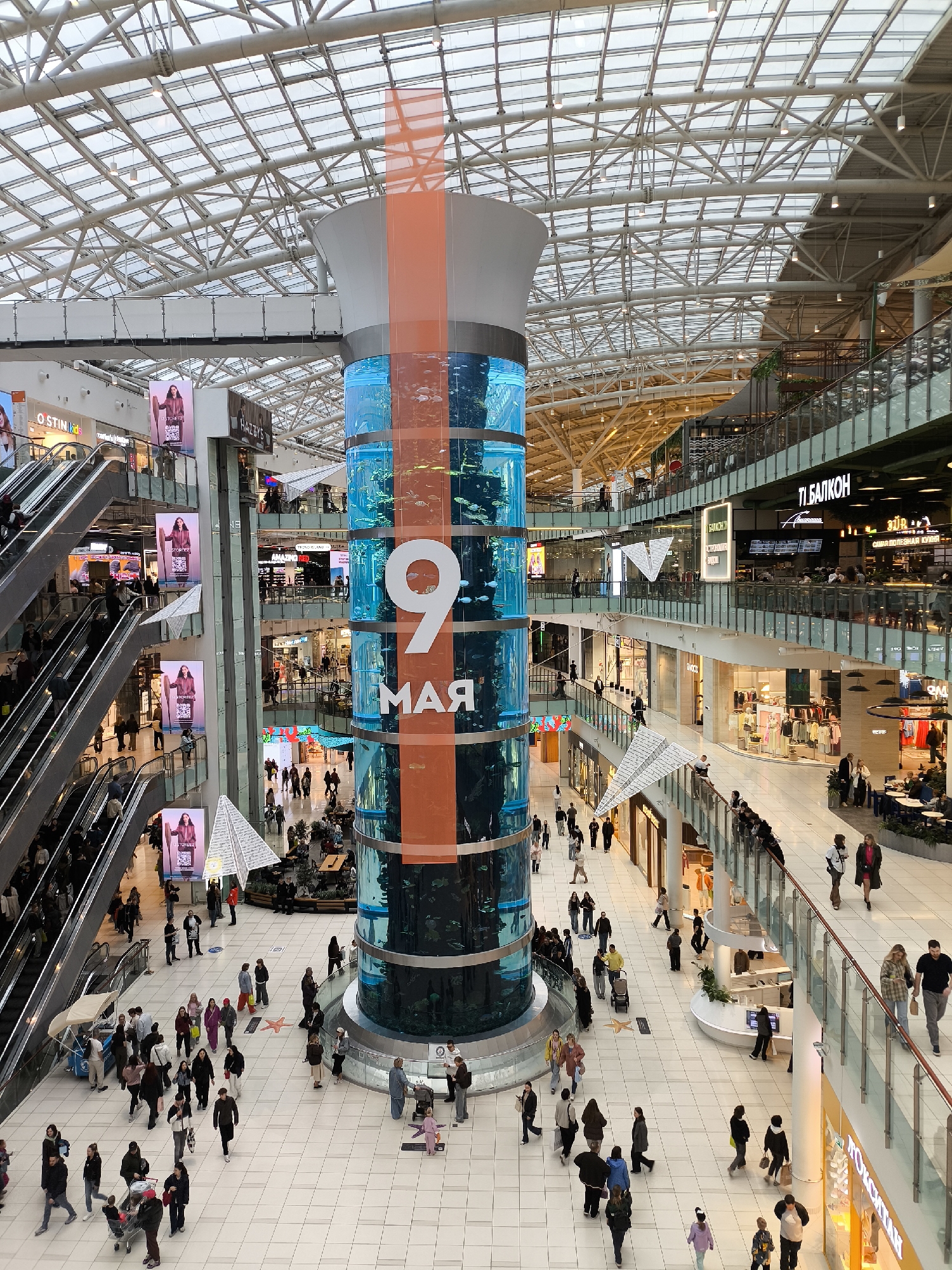 This is an indoor view of a modern shopping mall in Krasnogorsk, Russia, featuring a large cylindrical aquarium prominently displayed in the center. The aquarium is adorned with an orange banner displaying the text "9 МАЯ", likely referencing Victory Day. The mall has multiple levels, accessible via escalators. Numerous people are walking and browsing in the mall. Storefronts, restaurants, and advertisements are visible throughout the mall. The ceiling is a grid-like structure with skylights.