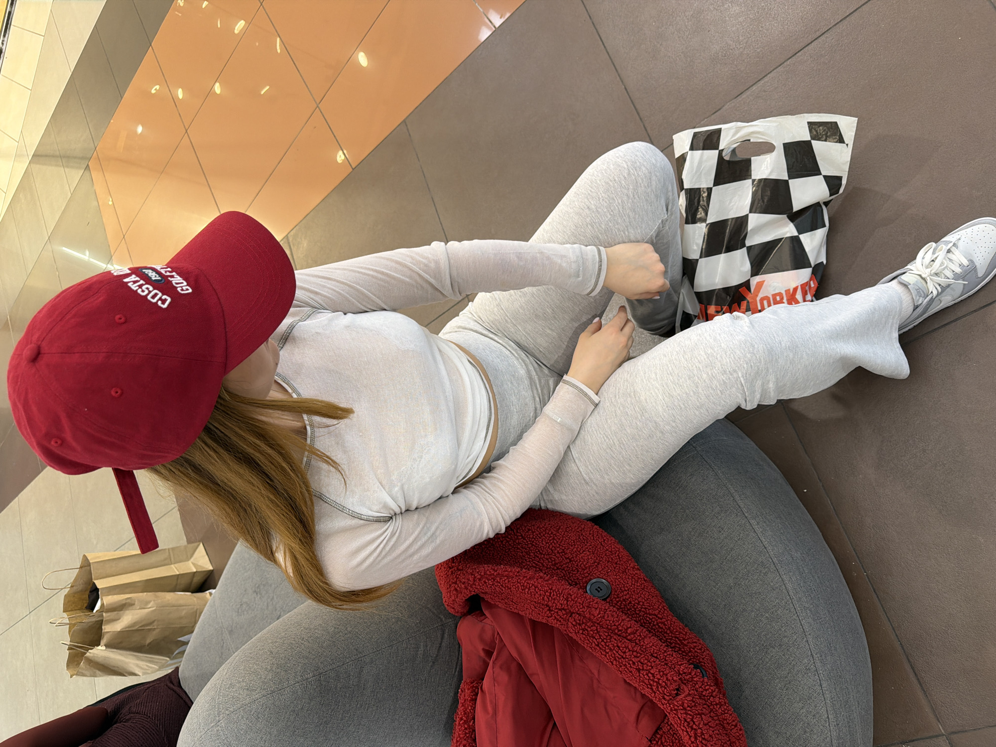 A woman wearing a red baseball cap with white text is sitting on a grey beanbag chair. She is wearing a long-sleeved light grey top and matching sweatpants, with white sneakers. To her right is a black-and-white checkered shopping bag with "New Yorker" printed on it. To her left are two brown paper shopping bags. A red jacket with a sherpa-like texture and a red lining is draped over the beanbag chair. The floor is tiled in orange and brown. The location is likely a shopping mall, but the exact location within Sofia, Bulgaria is not identifiable from the image.