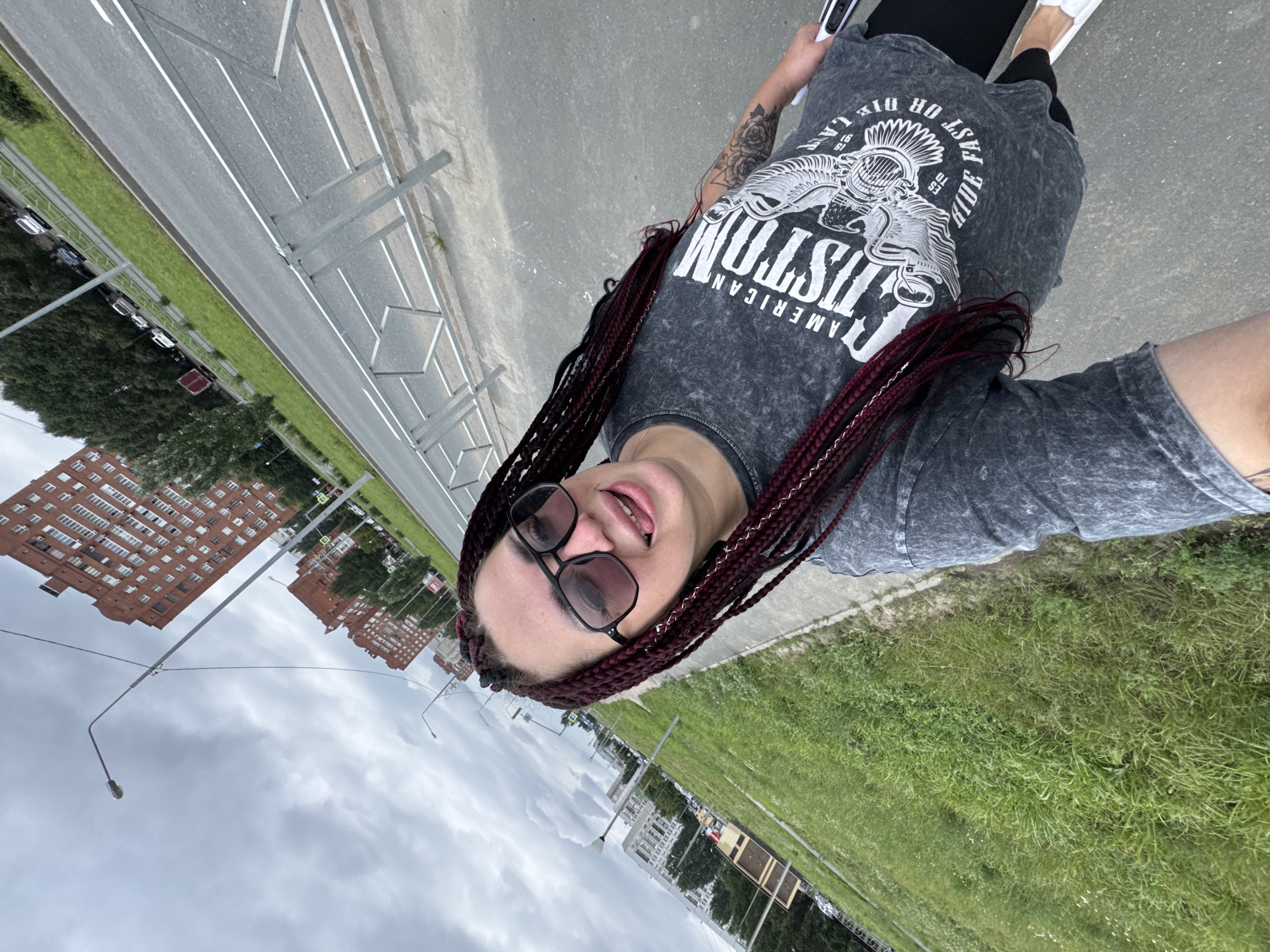 A woman with dark braided hair and sunglasses sticks her tongue out at the camera. She is wearing a grey t-shirt with a graphic print and black leggings. She is standing on a sidewalk next to a grassy area and a road. In the background, there are several apartment buildings, trees, and streetlights. The sky is cloudy. The image was taken in the city of Tolyatti, Russia.