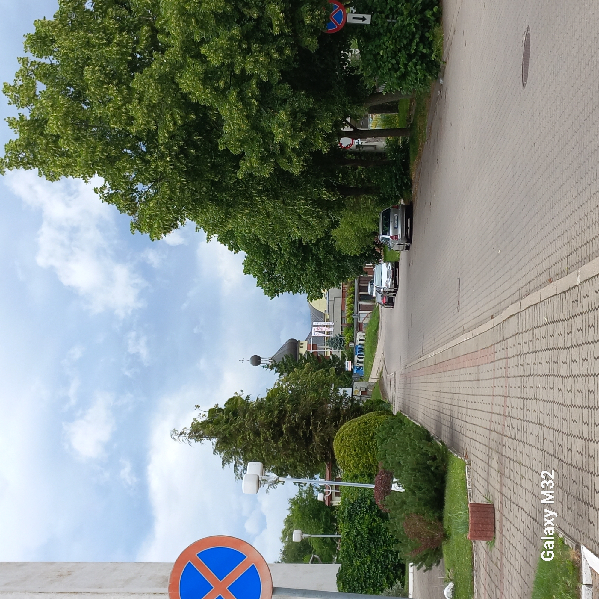 The image depicts a residential street in Olsztyn, Poland, on a cloudy day. A paved walkway lines the left side of the street, bordered by manicured bushes and grass. A traffic sign prohibiting parking is visible on the left. Several cars are parked along the right side of the road, which is also paved with interlocking bricks. In the background, a building with a domed roof, possibly a church, can be seen, surrounded by trees and other structures. The overall impression is one of a quiet, well-maintained urban environment.