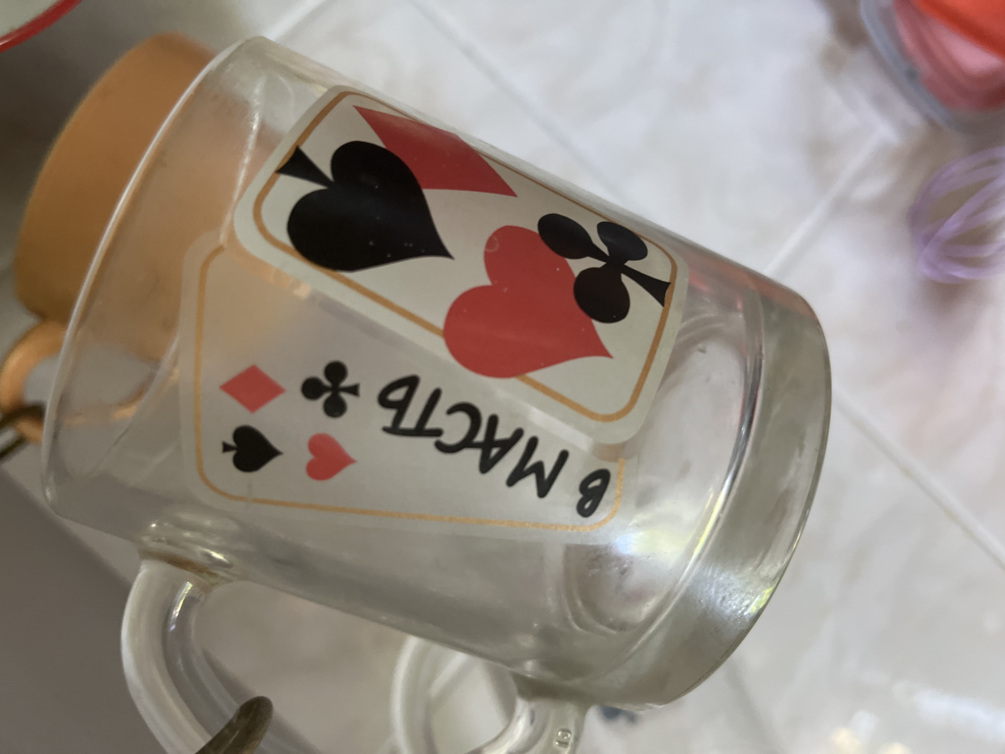 A close-up, eye-level shot shows a transparent glass mug decorated with playing card motifs. The mug is adorned with a diamond, spade, and heart symbol, along with the Russian text "в масть." The handle of the mug is visible on the left side, and a portion of an orange ceramic object is seen behind it. The background is blurred, with hints of a tiled surface and some colorful objects, possibly in a kitchen or storage area. There are no people present in the image. The image was taken indoors, with artificial lighting. There is no indication that the image is located in Minsk, Belarus, or any other specific city.