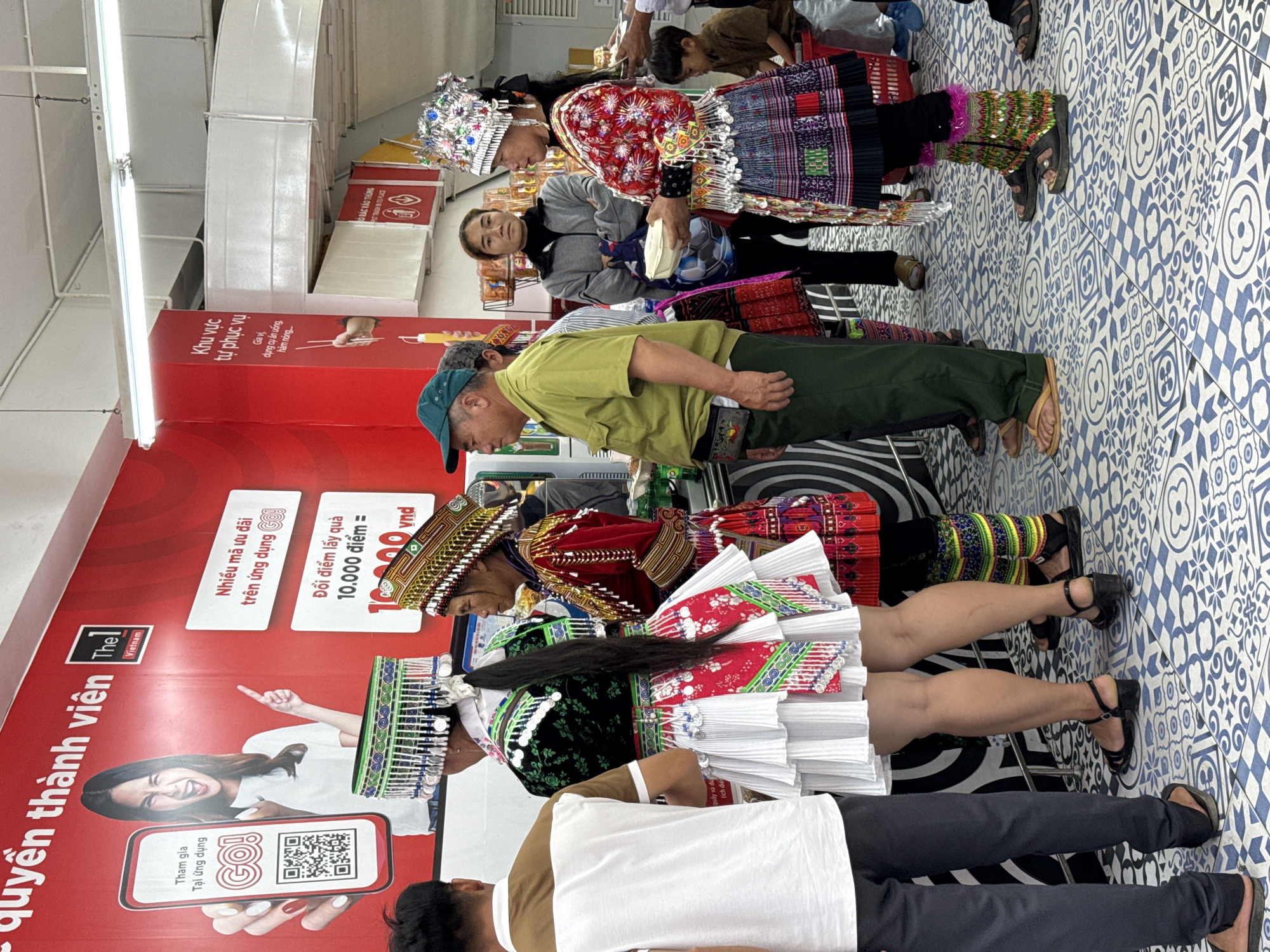 The image shows several people standing in a supermarket aisle, likely in Da Lat, Vietnam, based on the signage. Two women in traditional Hmong attire, including headdresses and colorful skirts, are near a red wall with Vietnamese text and branding for "Thel Vietnam" and "GO!" along with a QR code. A man in a green shirt and cap stands near the women, and other individuals are partially visible in the background. The floor has a patterned blue and white tile design. The supermarket features include displays advertising offers and a self-service area.
