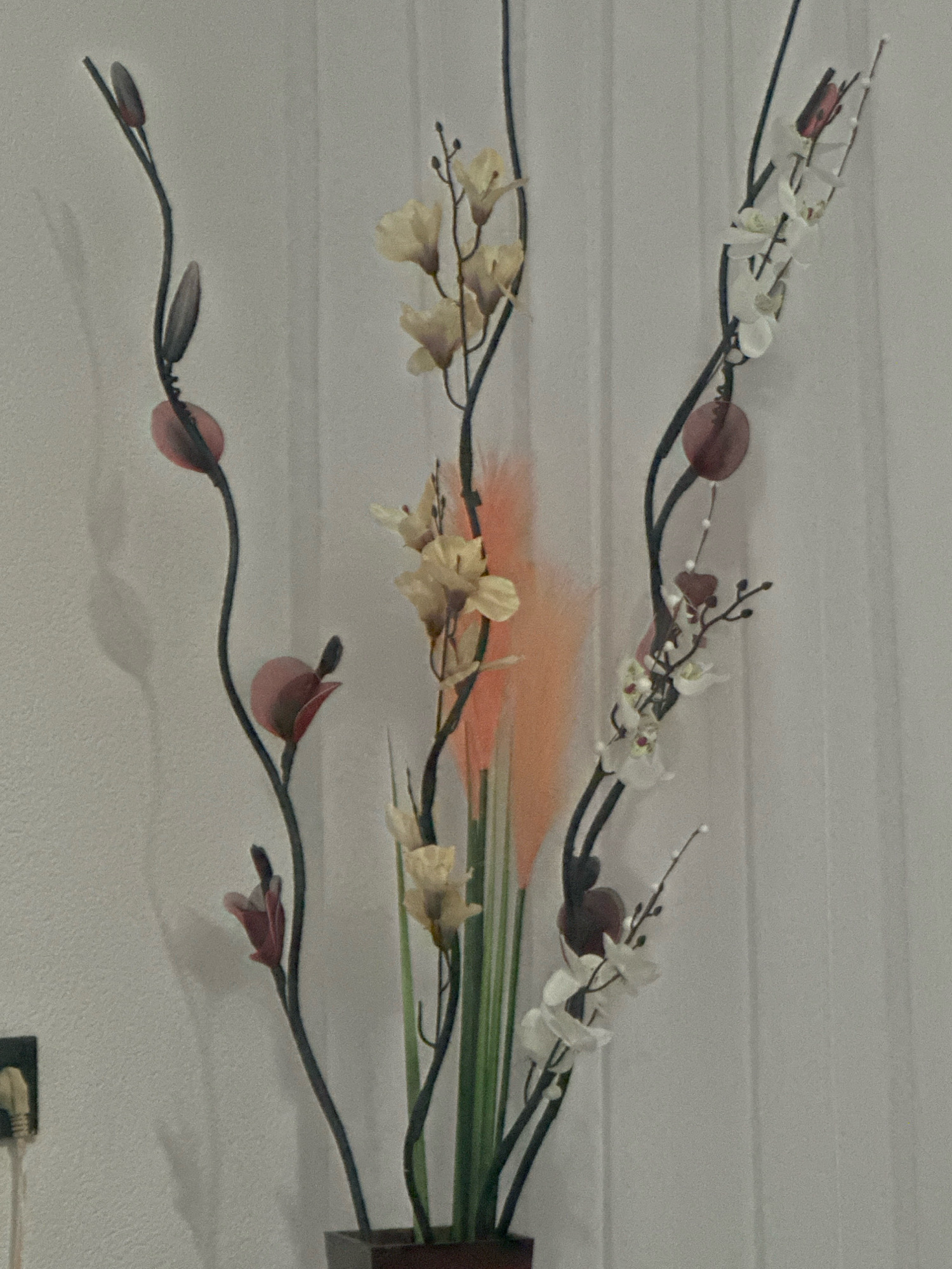 The image displays a close-up of an artificial floral arrangement set against a lightly textured white wall with vertical paneled blinds on the right. The arrangement features several dark, slender stems adorned with various artificial elements. On the left, there are dark, elongated buds or leaves and circular reddish-pink beads. Towards the center, stems hold clusters of cream-colored flowers and wispy, orange-pink feathery plumes. On the right, a stem is decorated with small white orchid-like flowers, a reddish-brown heart-shaped ornament, and more circular reddish-pink beads. The arrangement is housed in a dark rectangular vase, mostly obscured from view. The lighting casts soft shadows of the stems and leaves onto the wall. The scene is indoors, in a domestic setting. The location of Llukac i Begut, Kosovo, is not discernible from the visual information provided in the image.