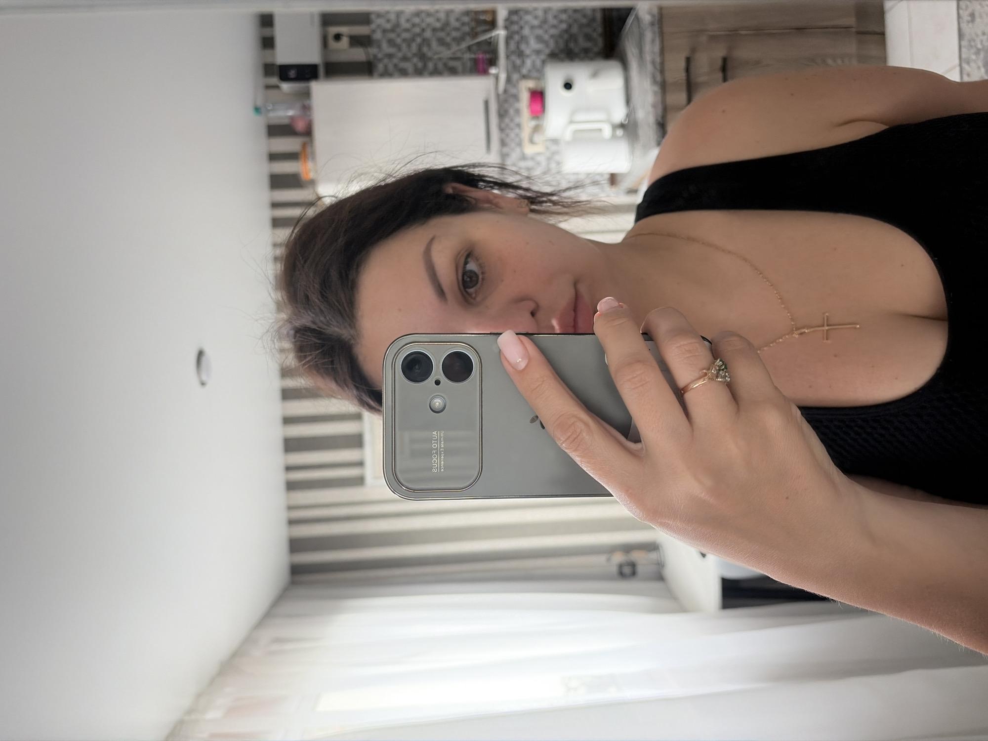 A woman is taking a mirror selfie in what appears to be a domestic setting, possibly a kitchen or bathroom. She is holding a silver smartphone in her left hand. The woman has dark brown hair, pulled back from her face, and is wearing a black tank top. She is also wearing a gold necklace with a cross pendant and a ring on her left hand with green stones. The background shows kitchen cabinets, a microwave, and a window with sheer curtains. There are no visible signs that this location is in Sochi, Russia.