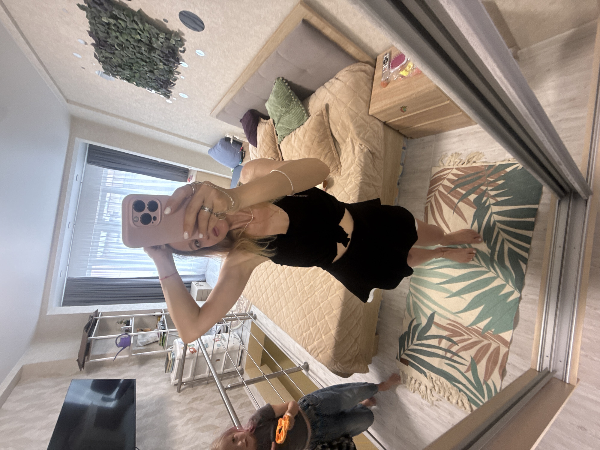A young woman with blonde hair is taking a mirror selfie in a bedroom. She is wearing a black crop top and a black skirt. Her hands are on her head and she is holding a pink phone. In the background, a child with pink hair is standing near a railing. The room has a bed with light brown quilted bedding, a wooden nightstand, and a rug with a tropical leaf pattern. A television is mounted on the wall.