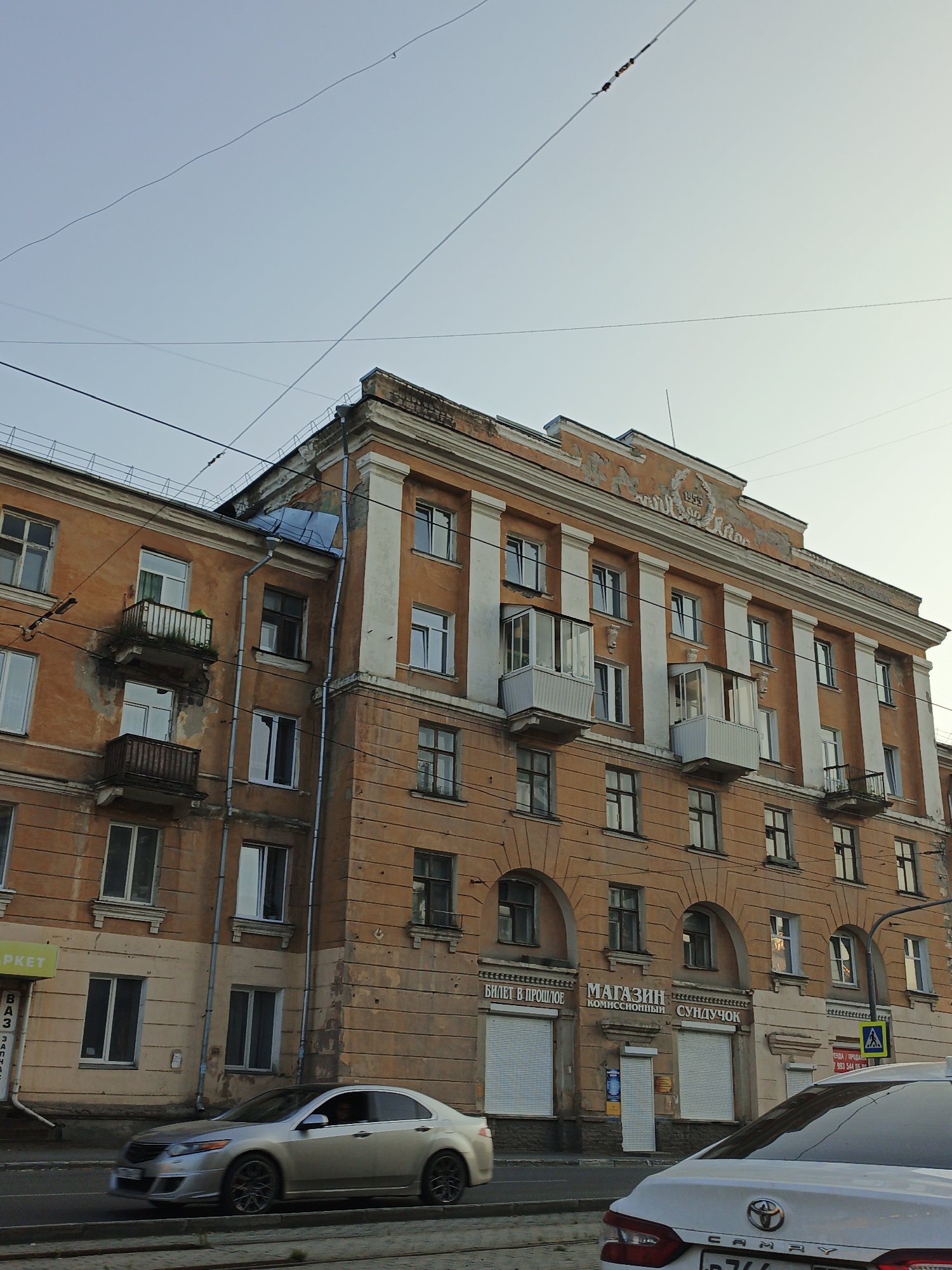 The image shows a multi-story building in Zlatoust, Russia. The building is orange with white trim and columns. There are many windows and several balconies. Some of the paint on the building is peeling. There are power lines running across the sky in front of the building. In the foreground, a silver car is driving on the road. The sky is clear and the sun appears to be low in the sky, suggesting it is either early morning or late afternoon. The building has signage in Russian. One sign reads "МАГАЗИН КОМИССИОННЫЙ СУНДУЧОК" which translates to "Consignment Store Chest". Another sign reads "БИЛЕТ В ПРОШЛОЕ" which translates to "Ticket to the Past". There is a yellow traffic sign with a blue pedestrian symbol.