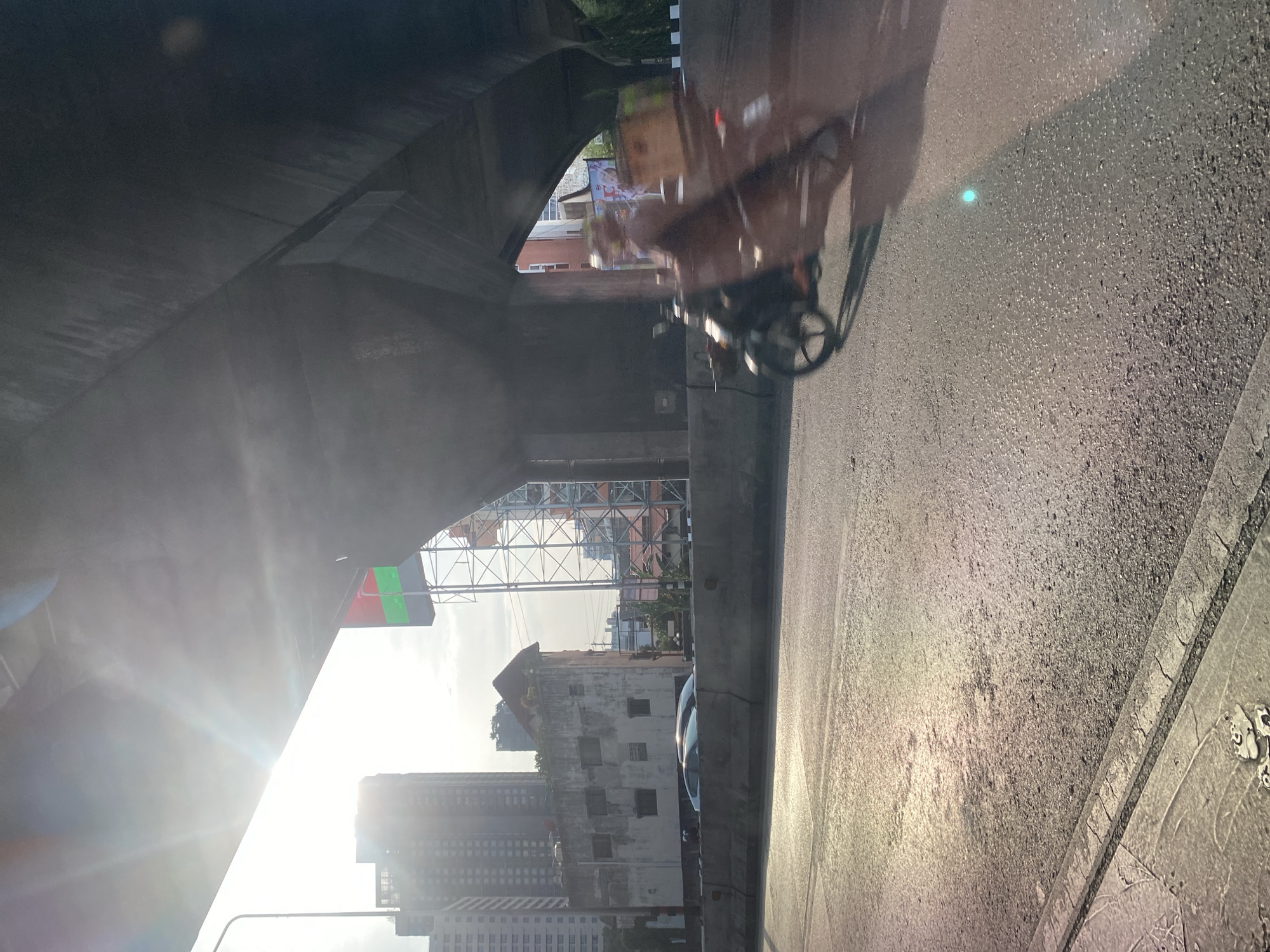 The image depicts a street scene in Bangkok, Thailand, captured from a low angle perspective, likely from a vehicle. Overhead, a large concrete structure, possibly an elevated roadway or bridge, dominates the upper left quadrant. Sunlight streams into the scene, creating glare and obscuring details.
A three-wheeled vehicle, likely a motorbike taxi or a delivery cart, is positioned on the right side of the frame. The surrounding environment includes buildings and infrastructure typical of a dense urban setting. High-rise buildings are visible in the distance, along with a large billboard structure. The street surface appears to be asphalt, showing wear and tear.