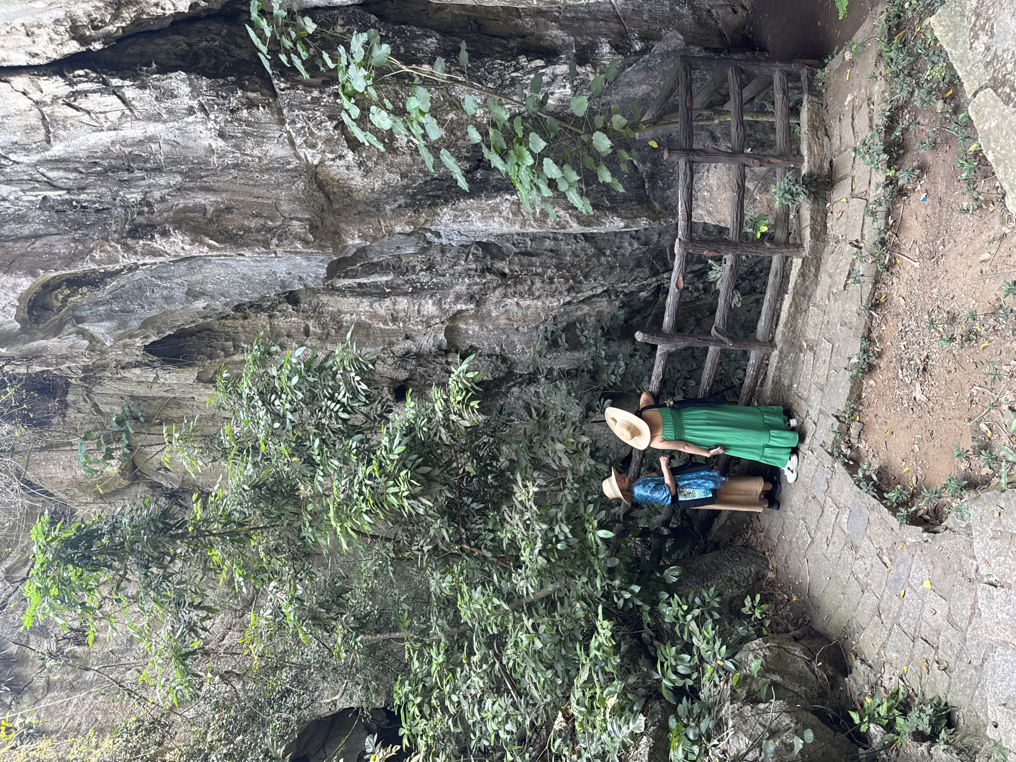 Two women wearing hats stand on a stone pathway at the Marble Mountains in Đà Nẵng, Vietnam. One woman wears a green dress, and the other wears a blue shirt and tan skirt. A wooden fence borders the pathway. A cliff face is to the left, covered in vegetation.