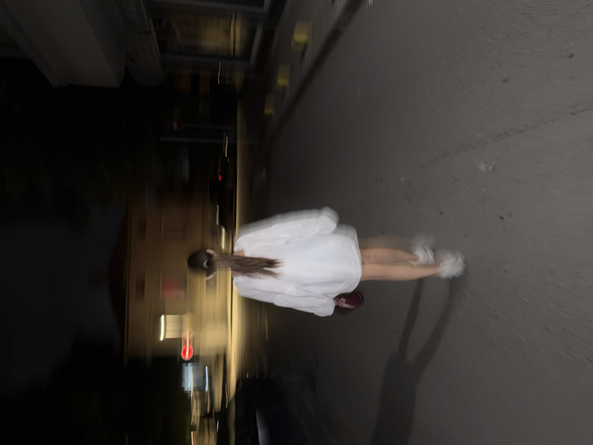 The image depicts a blurry, low-light scene at night in an urban environment, likely a street or alleyway in Plovdiv, Bulgaria. The primary subject is a person, appearing female, viewed from the back. She has dark hair tied in a ponytail and is wearing a light-colored, possibly white, long-sleeved top or dress that extends to mid-thigh, paired with light-colored shoes, possibly sneakers. One foot has a darker shoe (reddish-brown).
The person appears to be in motion or captured with significant camera shake or a long exposure, resulting in a pronounced motion blur across the entire frame. She is positioned on a paved surface, likely asphalt or concrete, which covers the right two-thirds of the image. Long, dark shadows are cast on the ground to her right.
In the background, on the left side of the frame, are dark, indistinct buildings. Streaks of blurred light, indicative of illuminated windows or streetlights, are visible against the dark structures, further emphasizing the nighttime setting and motion blur. No specific activities or events are clearly discernible beyond the presence of the person on the street. There is no visible text. The overall atmosphere is dim and indistinct due to the low light and blur.