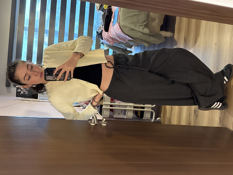 A young woman takes a mirror selfie. She wears a black crop top, loose grey drawstring pants, a cream-colored cardigan, and black Adidas sneakers. Her hair is pulled back, and she wears earrings and a bracelet. The background includes a window with blinds, a bed with a green comforter, a metal rack, and a door. The image is slightly tilted.