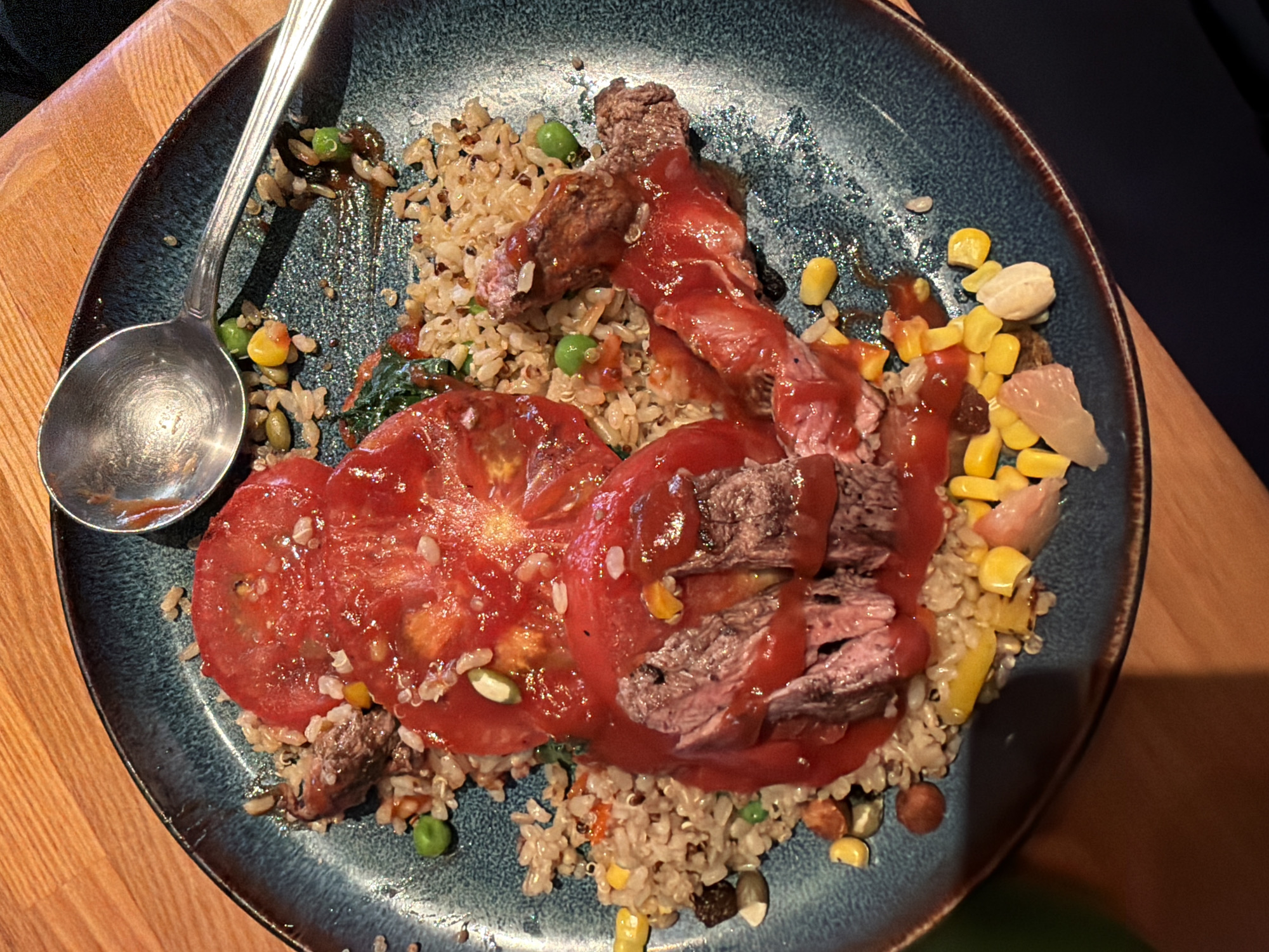 A close-up, overhead shot shows a plate of food on a wooden table. The plate is dark blue with a brown rim and contains a dish of rice, corn, peas, and meat topped with sliced tomatoes and a red sauce. A spoon rests on the edge of the plate. The lighting is warm and the image is slightly blurry. The location of Yuzhong District, China is not visible in the image.
