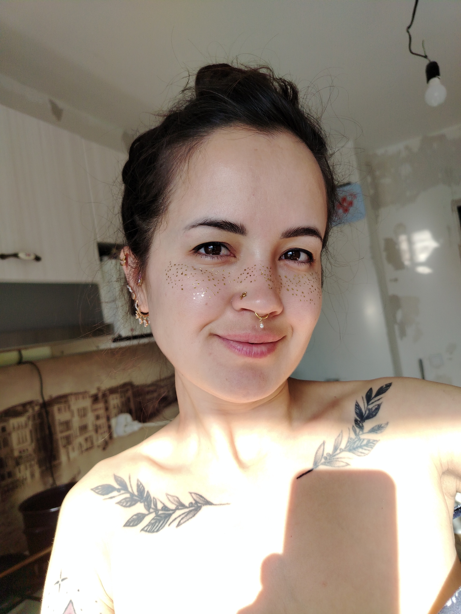 In a close-up, indoor shot, a woman with dark hair pulled up in a bun smiles at the camera. She has gold glitter applied to her cheeks, mimicking freckles. She is also wearing a nose ring with a small pearl and earrings with dangling charms. Tattoos are visible on her upper chest and shoulder, featuring a branch with leaves. The background appears to be a room under renovation, with unfinished walls and a hanging light fixture. The lighting suggests it is daytime, with sunlight streaming in from the right. The woman is positioned in the foreground, with the environment blurred behind her. There is no specific indication of the location being in Noviy, Russia.