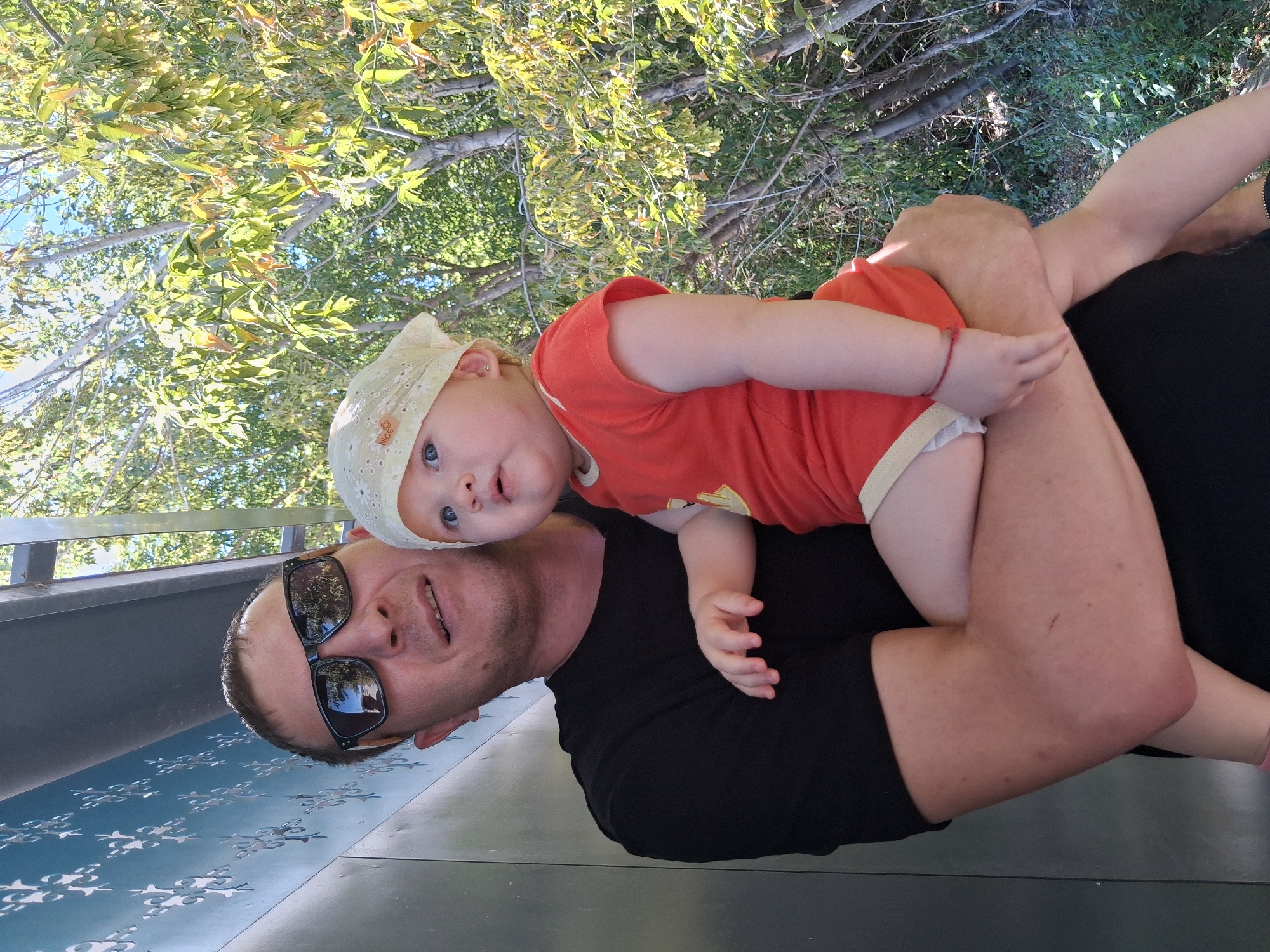 A man wearing sunglasses and a black t-shirt holds a baby dressed in an orange shirt and a white bandana. The man's left arm is around the baby's waist, and his right arm is supporting the baby's legs. The baby looks to its left, with wide eyes. They are outdoors, with a textured blue wall on the left and trees with green leaves in the background. A hint of a grey overhang is visible above. The image is taken in Semey, Kazakhstan.
