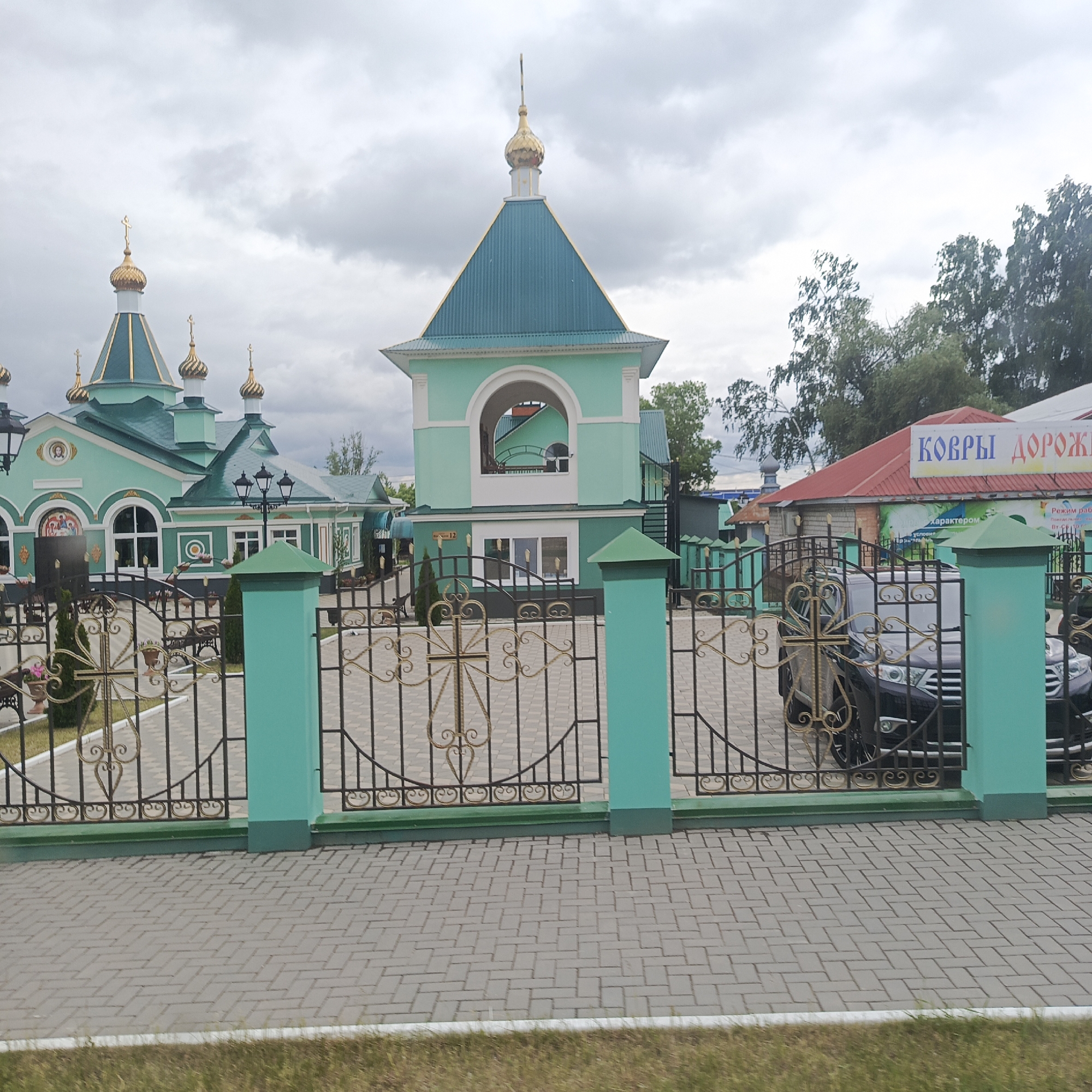 The image captures an outdoor scene in Chaikovsky, Russia, dominated by a complex of buildings exhibiting architecture typical of Russian Orthodox churches. The main structures are painted in shades of teal and light green, featuring dark teal roofs and numerous golden onion domes, each topped with a cross. A prominent bell tower-like building in the center has a large archway and a golden spire.
In the foreground, a gray paved area with interlocking bricks in a herringbone pattern leads to a decorative wrought iron fence. The fence is black with ornate gold scrollwork, and its teal support pillars match the buildings. Notably, one section of the fence gate incorporates a golden cross design.
Behind the fence, a dark SUV is partially visible on the right, parked near a building with a contrasting red corrugated roof. A large sign on this red-roofed building clearly displays "КОВРЫ ДОРОЖИ" (Kovry Dorozhi), likely indicating a shop selling carpets or rugs. Another sign below it shows "характером" (character). A small white sign with "№ 12" is affixed to the teal building with the archway.
The sky is overcast with gray clouds, suggesting a cloudy daytime. Green grass borders the paved area in the immediate foreground, and lush green trees are visible in the background on the right. The overall atmosphere is tranquil, with no people visible in the frame.