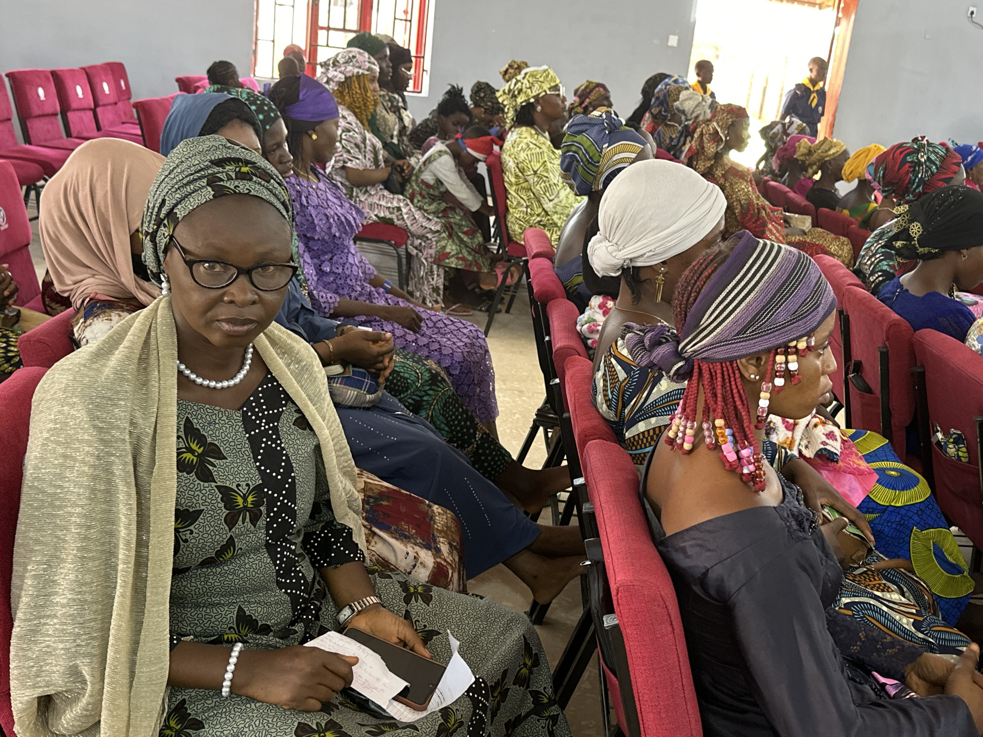 This image captures an indoor gathering of approximately 30-40 individuals, primarily women, seated in rows of red upholstered folding chairs within a room in Goni Gara, Nigeria.
In the foreground, a woman with dark skin and dark-framed glasses looks directly at the camera with a neutral expression. She wears a patterned headwrap in shades of light green and black, a light green dress with black butterfly and white dot patterns, and a light gold-colored shawl. She is adorned with a pearl necklace and a pearl bracelet on her left wrist. Her left hand rests on her lap, holding a black smartphone and a folded piece of white paper with visible writing.
To her immediate right, a woman's profile is visible, featuring dark skin and reddish braided hair adorned with numerous colorful beads. She wears a dark long-sleeved top and is seated facing away from the camera, towards the right.
Behind the foreground woman, multiple other women are seated, predominantly wearing vibrant, patterned fabrics and headwraps. Their attire showcases a variety of colors and designs, including geometric, floral, and abstract motifs. One woman behind the foreground subject has a peach-toned head covering, and another further back wears a blue head covering. Many individuals appear attentive, some looking forward, others turned slightly.
The room features light grey or off-white walls. On the left side of the background, several unoccupied red chairs are visible, alongside a window with red frames. In the far background, near a brightly lit area that suggests an open doorway or large window, two young individuals are visible, one wearing a yellow and blue uniform. The floor appears to be a light-colored, possibly concrete or tiled surface. Natural light illuminates the scene from the background.