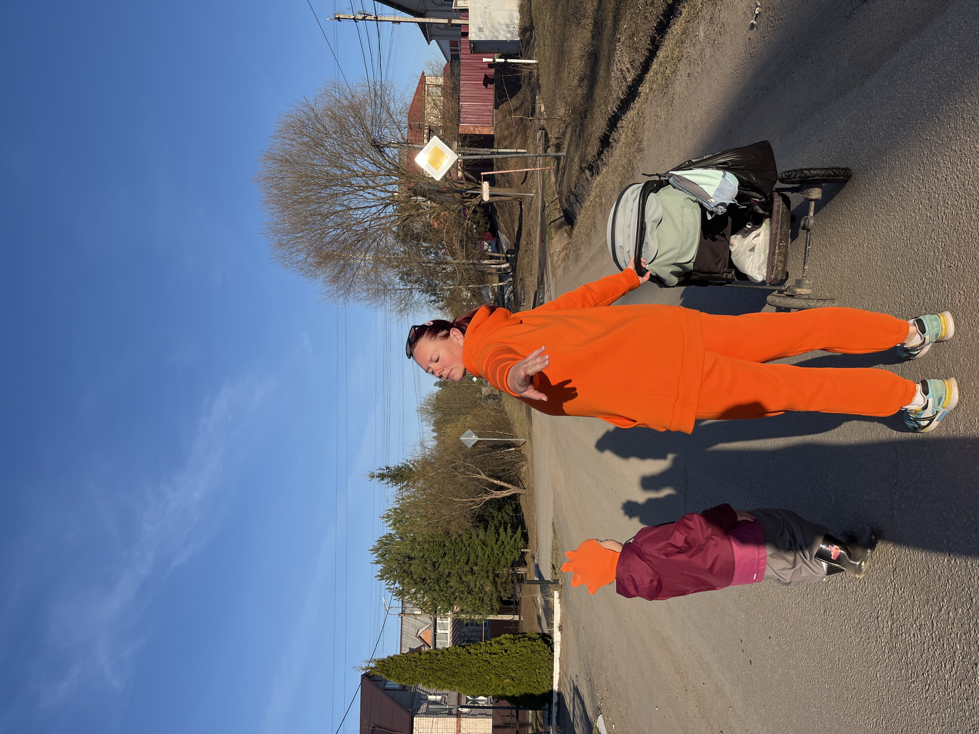 An outdoor, rotated full shot captures a woman and a child on a paved road in what appears to be a semi-rural or village setting. The woman, in an bright orange tracksuit and sneakers, stands with her arms slightly outstretched, facing towards the left of the frame. Her brown hair is tied back, and she wears sunglasses on top of her head. To her right, a child is partially visible, wearing a maroon jacket, grey pants, and an orange hat, standing near her feet.
Beside the woman, a grey utility cart or modified stroller, laden with bags and possibly other belongings, suggests travel or transport. The road has a dirt shoulder on both sides, and in the background, there are several houses, some bare deciduous trees, and some evergreen trees under a clear blue sky with sparse clouds. Utility poles with wires run along the side of the road. A yellow diamond-shaped road sign, indicating a main road or right-of-way, is visible further down the road. The bright sunlight casts distinct shadows of the woman and child on the asphalt, suggesting either early morning or late afternoon. The overall scene depicts a calm, sunny day in a small community in Chaikovsky, Russia. No clearly legible text is visible.