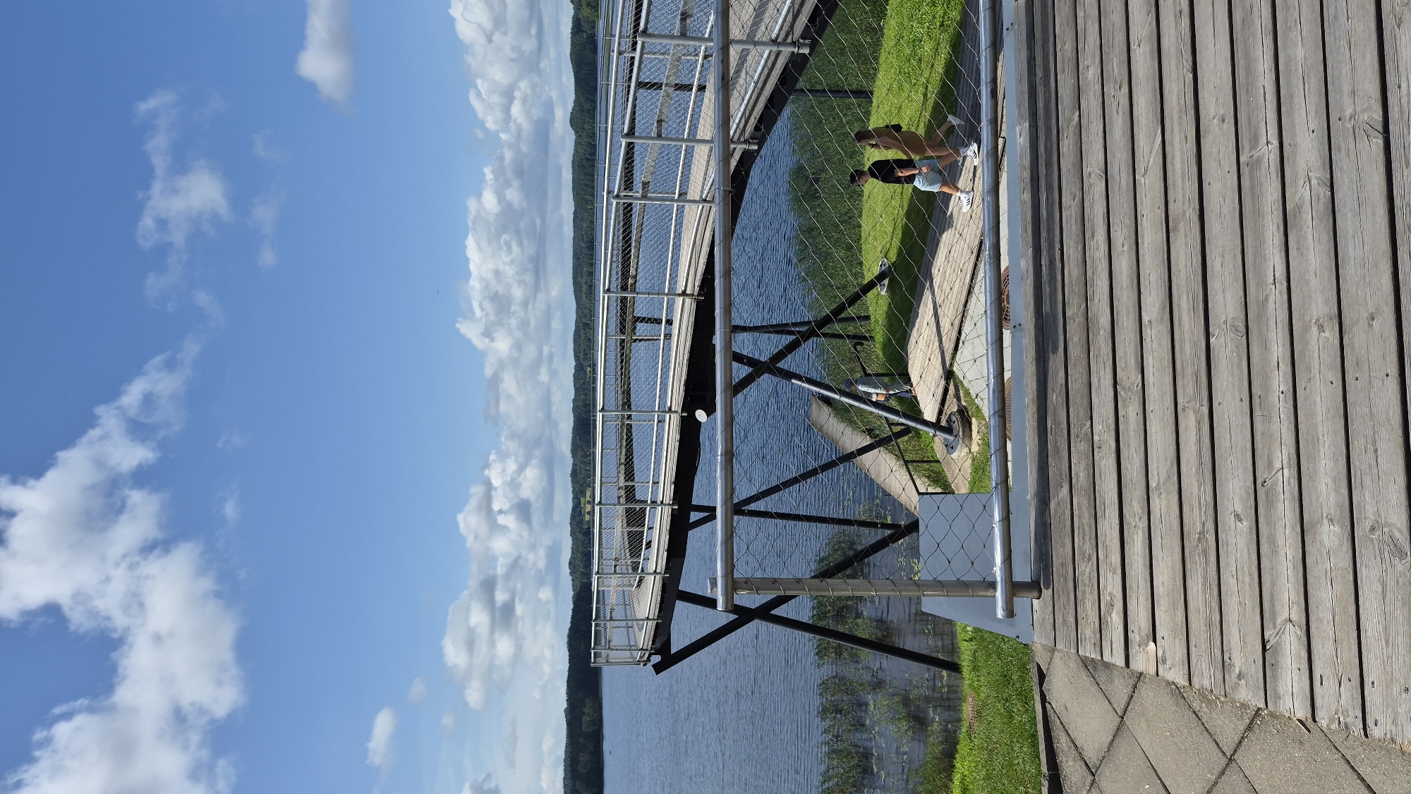 An elevated wooden walkway with metal railings curves over a calm blue lake under a bright sky with scattered white clouds. The walkway is supported by black X-shaped metal stilts. On the grassy bank near the walkway, three people are visible: two young adults walking together, and one person standing further down the path. The foreground features a portion of a lower wooden deck. The background shows a treed shoreline and rolling hills. This scene depicts the Zarasai Skywalk in Zarasai, Lithuania, a popular tourist attraction offering panoramic views of the lake and surrounding landscape.