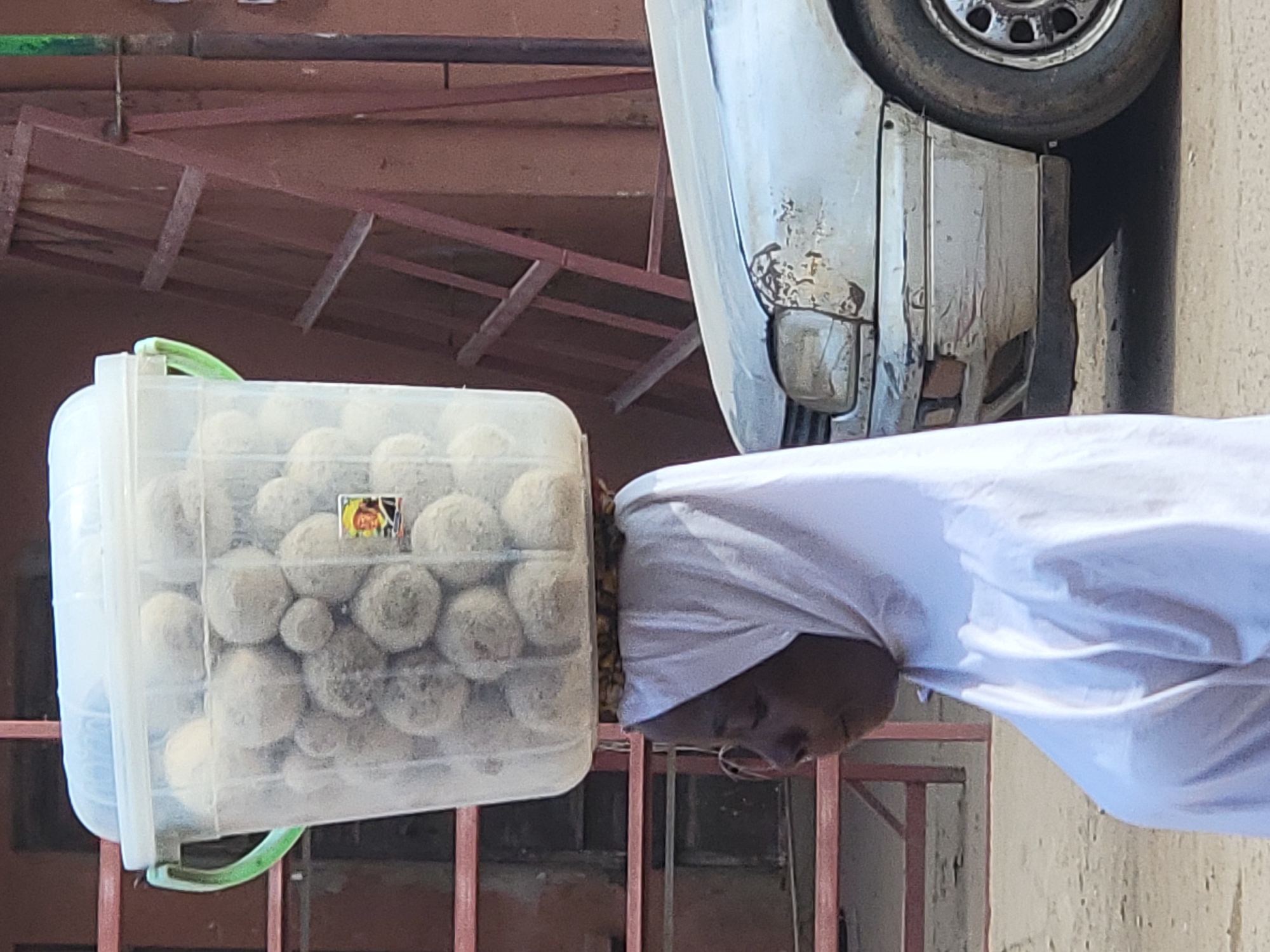 A person in a white garment carries a clear plastic container filled with round, textured objects on their head. The location appears to be an urban area, possibly Maiburiji, Nigeria. A damaged, light-colored vehicle with a visible tire and front quarter panel is parked nearby. The background shows architectural features, including a metal structure and a painted building with visible wear and tear. The scene is set outdoors in daylight.