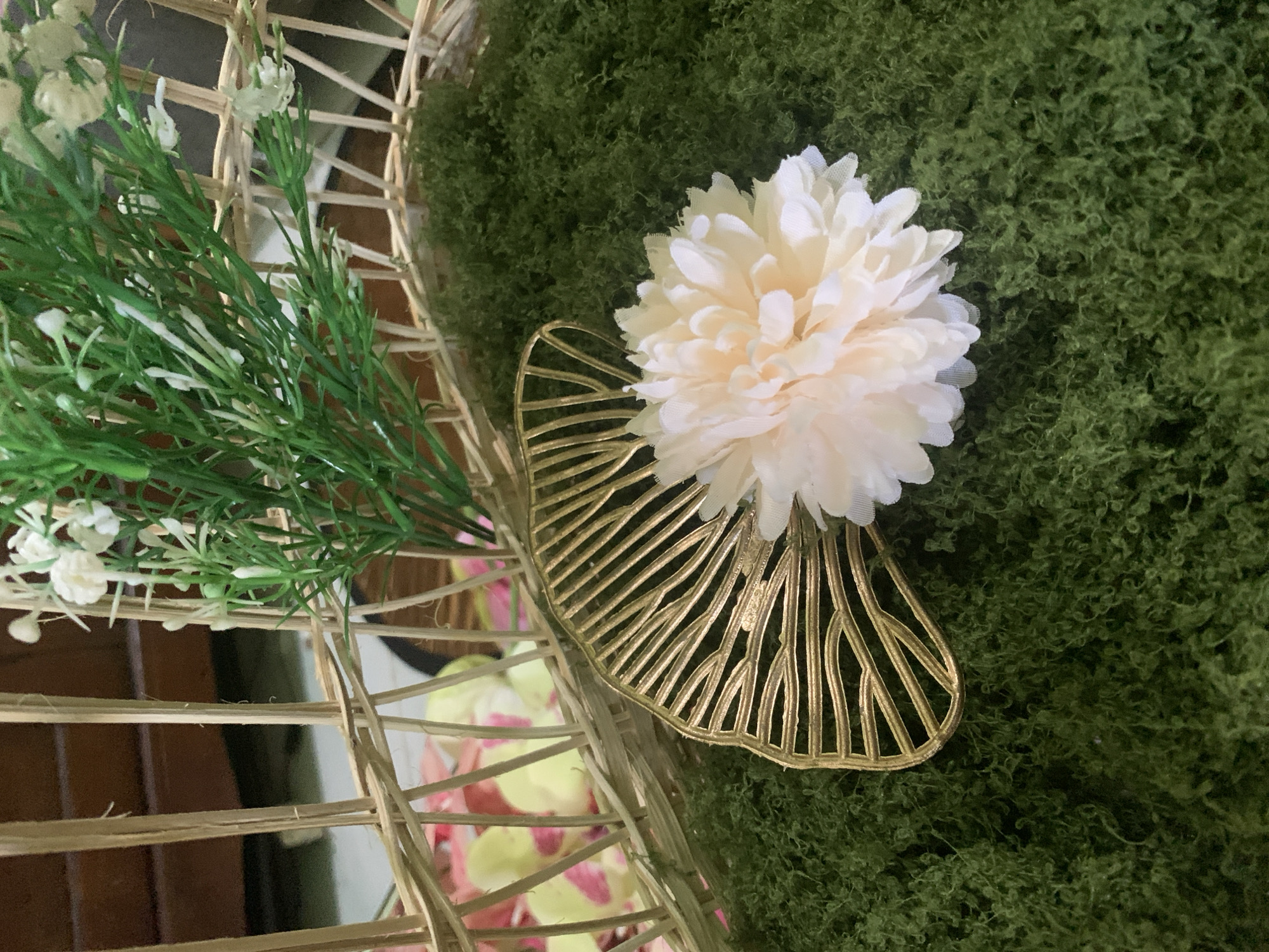 The image shows a close-up view of decorative elements placed within a wicker basket. The basket's woven structure is visible, with some areas covered in green, fluffy material resembling moss. A gold-colored, fan-shaped decorative piece with intricate cutouts is positioned on the moss. Resting on the gold piece is a light-colored, fluffy artificial flower, possibly peach or pale pink. Behind these elements, a sprig of artificial greenery with small white blossoms is also visible, protruding from the basket. In the background, a hint of a wooden structure and some scattered pink and yellow flower petals can be seen. The scene appears to be a still life arrangement, likely for decoration. There are no people or actions depicted in the image, and no specific location in Kuala Pembuang, Indonesia, can be identified from the visual information provided.