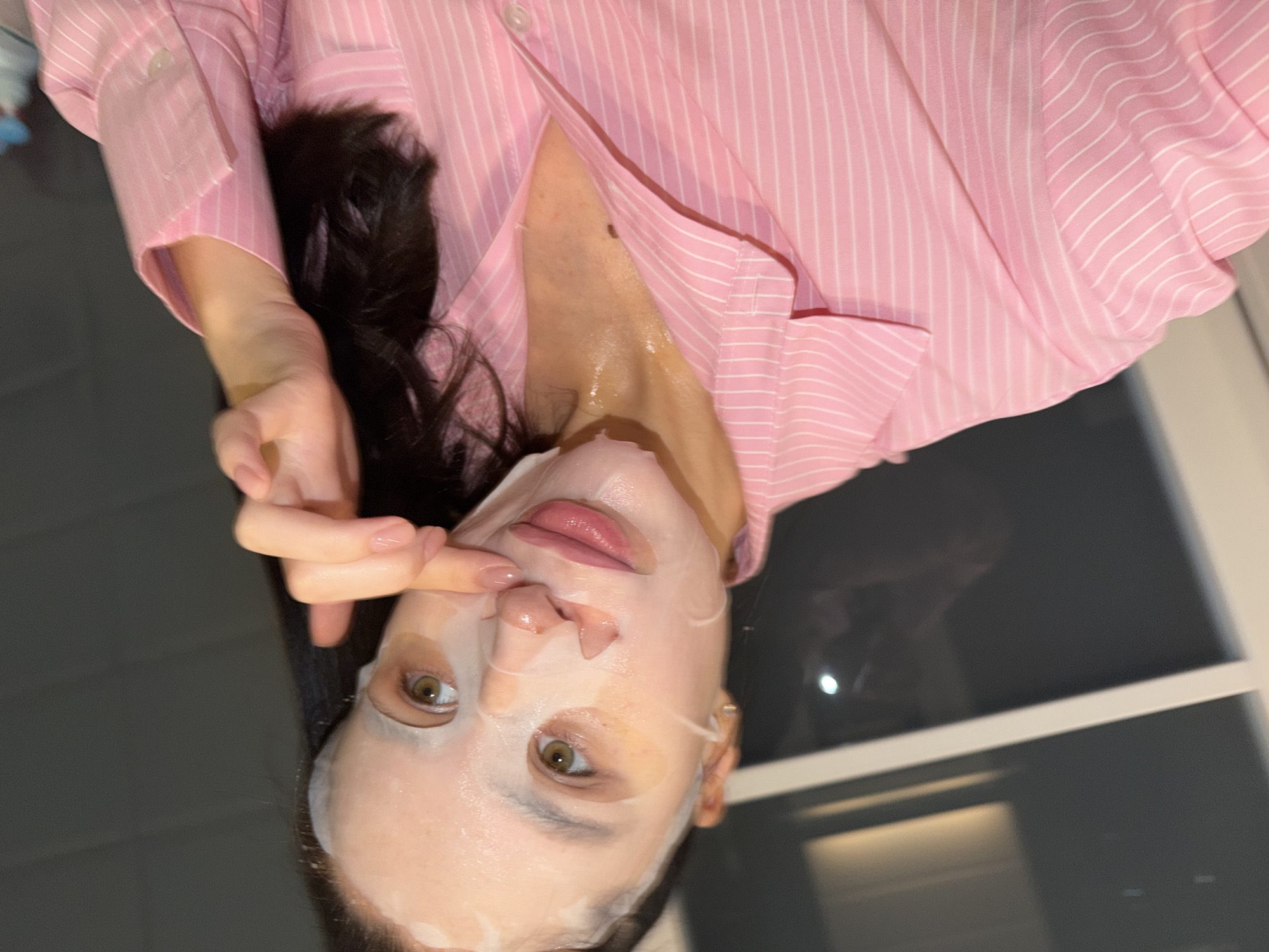 An upright portrait-oriented close-up shows a person, likely female, engaged in a self-care routine in Khimki, Russia. The individual has long, dark hair and is wearing a translucent sheet face mask that covers most of their face, leaving the eyes, nostrils, and part of the mouth exposed. Their eyes are open and appear to be light brown or hazel. One finger from their left hand, adorned with light-colored manicured nails, is gently touching the mask near the nose, seemingly to adjust or smooth it. The skin visible around the eyes and lips has a moist, dewy appearance from the mask.
The person is wearing a light pink button-down shirt with thin white vertical stripes. The collar and cuff of the shirt are visible, and the fabric appears soft and somewhat loose-fitting. The background is dark and indistinct, suggesting an indoor setting, possibly during the evening or night. On the right side, there are faint reflections of light sources on a dark, reflective surface, which could be a window or another furnishing. The overall impression conveyed is one of calm and personal indulgence. There is no visible text in the image.