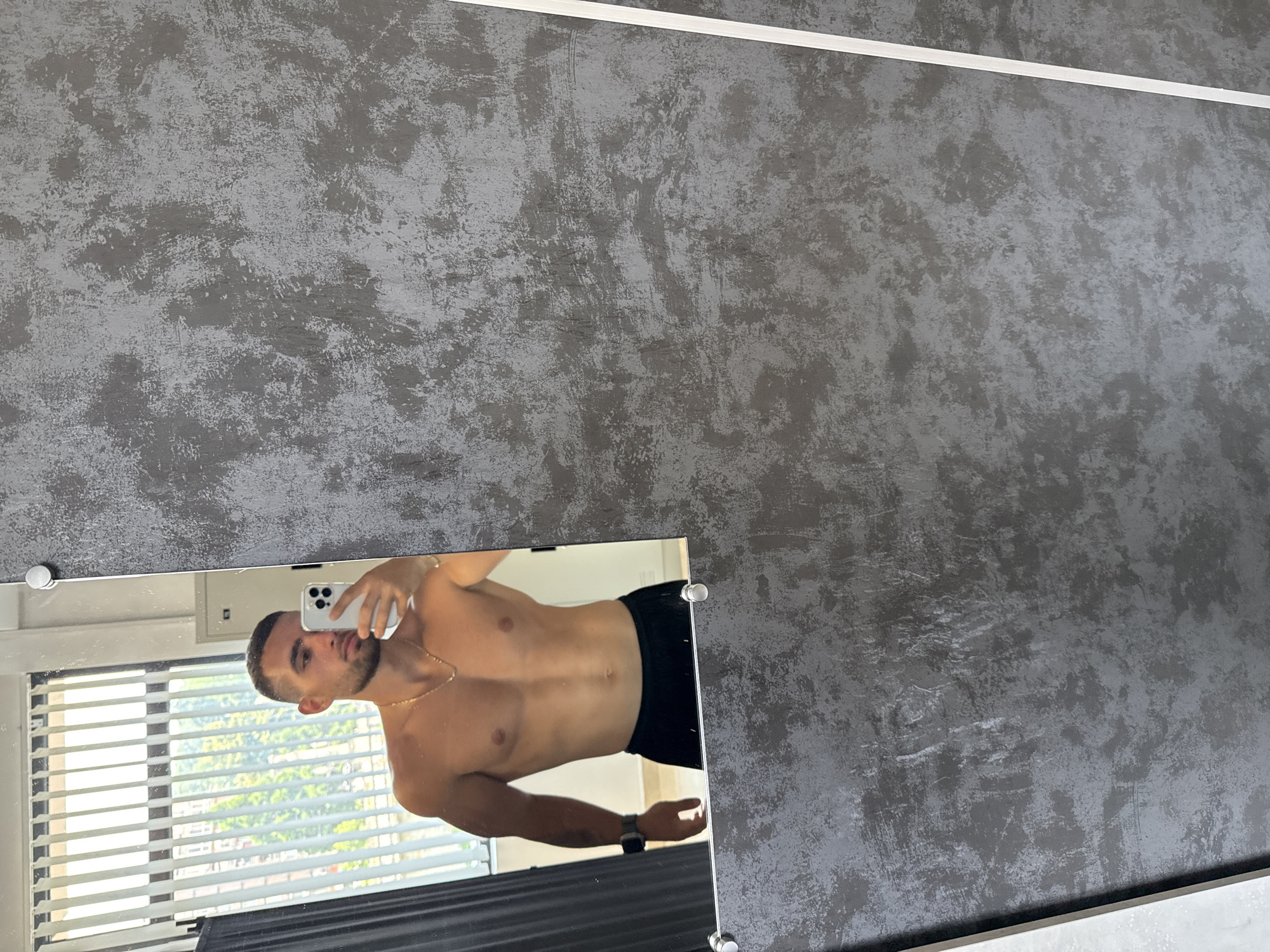 A shirtless man with tan skin and dark hair is taking a mirror selfie in what appears to be a gym locker room. He is wearing black shorts and a gold chain necklace. The mirror reflects vertical blinds and trees outside a window. The walls of the locker room are textured grey.