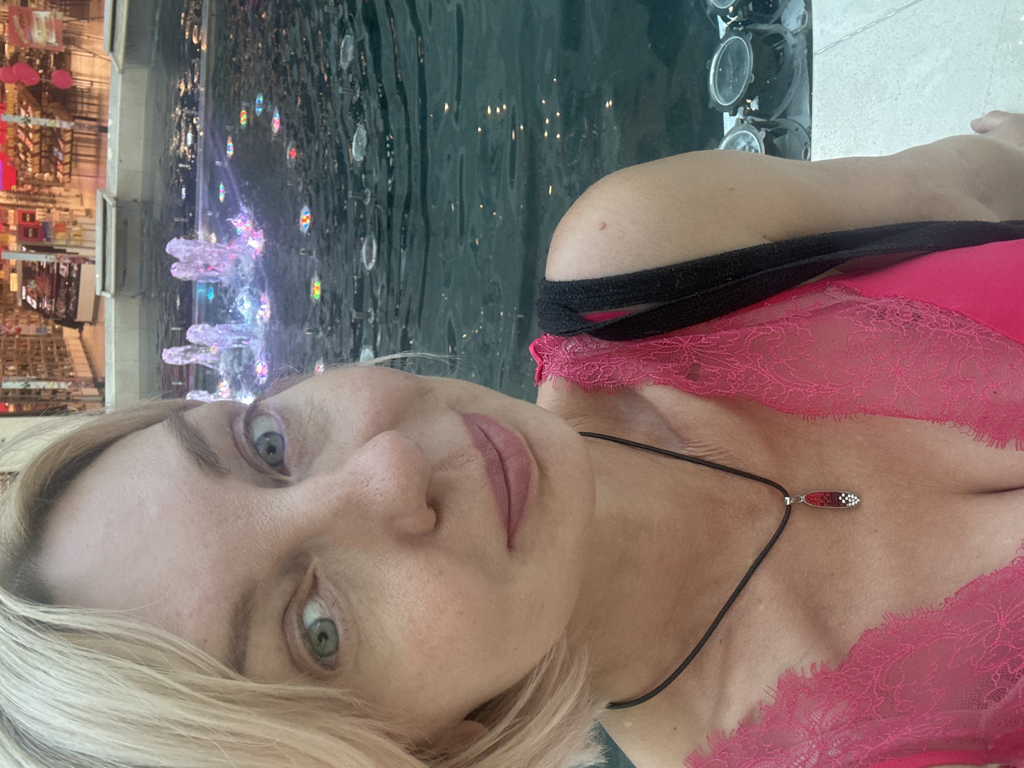 The image is a close-up, vertically oriented selfie of a fair-skinned woman with blonde hair and light green eyes. She is wearing a bright pink lace top and a dark cord necklace with a small, oval pendant. The pendant is silver with a red and white patterned inlay. The woman is looking directly at the camera with a neutral expression.
Behind her, a body of dark water is visible, with several colorful, illuminated jets of water creating a fountain effect. The water appears to be indoors, as suggested by the blurred background showing shelves filled with products and what look like artificial plants or decorations. The lighting is dim, with the fountain lights and the ambient lighting from the indoor setting creating reflections on the water's surface.
The overall scene suggests an indoor leisure area, possibly a shopping mall or hotel with a decorative fountain. The time of day is indeterminate due to the indoor setting. There are no visible text elements in the image. The woman's presentation and the lace clothing might suggest a relaxed or intimate atmosphere. The location context of Sochi, Russia, does not provide specific visual cues within the image itself.