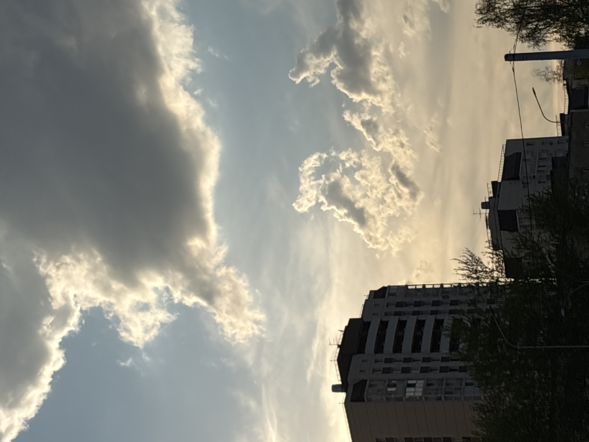 The image shows a cloudy sky in Perm, Russia. In the bottom right, there are several multi-story apartment buildings. Trees are visible in front of the buildings. A streetlight is also visible. The sky is light blue with several white and gray clouds.