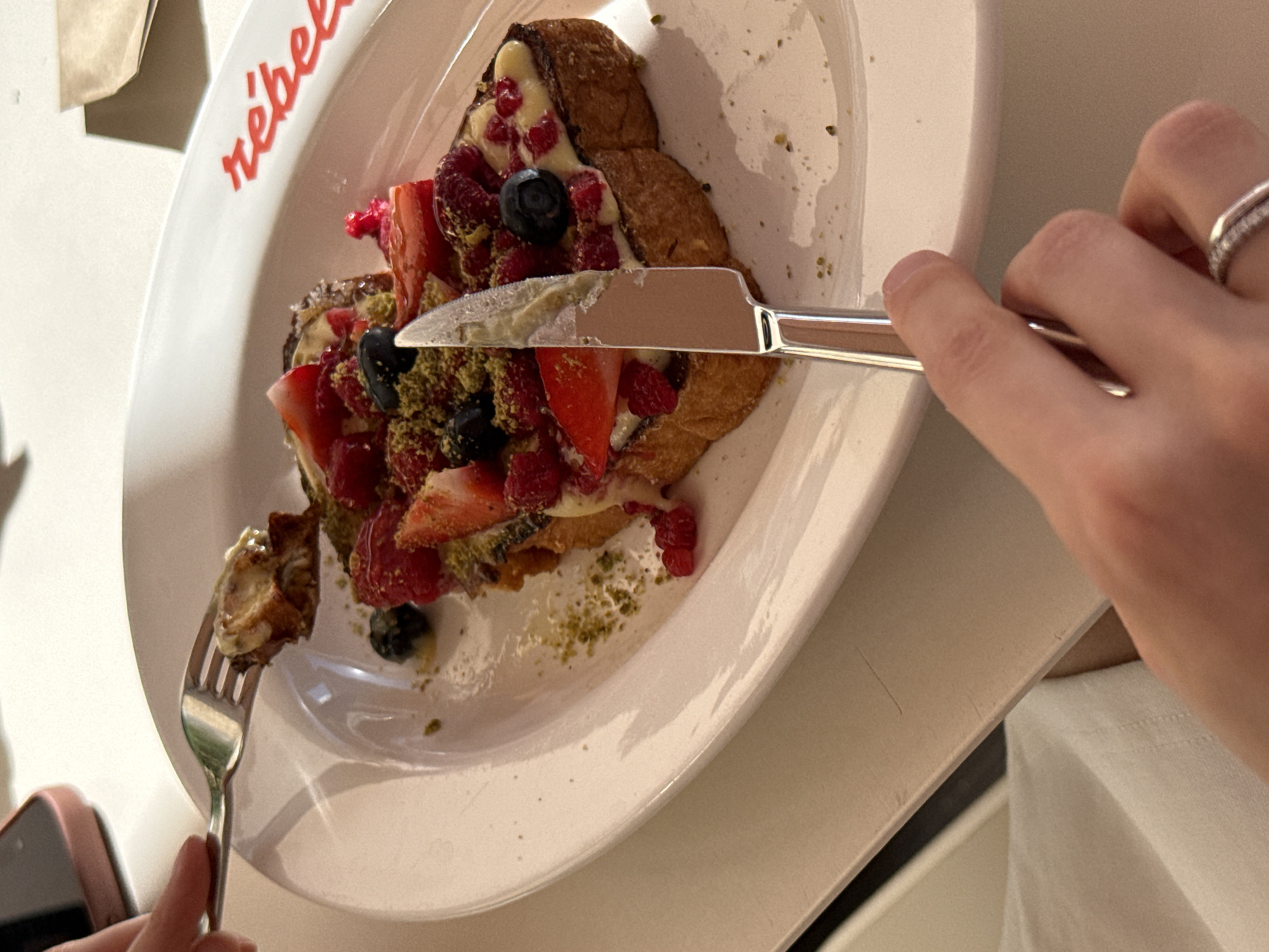 An overhead, medium shot captures a table setting with a plate of French toast topped with berries and pistachios. A fork holds a piece of the French toast, and a knife rests on the plate. The background is blurred, showing a portion of a pink mobile phone. The word "rebels" is visible in red lettering on the side of the plate. The scene suggests a meal in a restaurant, possibly in Moscow, Russia, given the branding on the plate which is reminiscent of a cafe that might exist there.