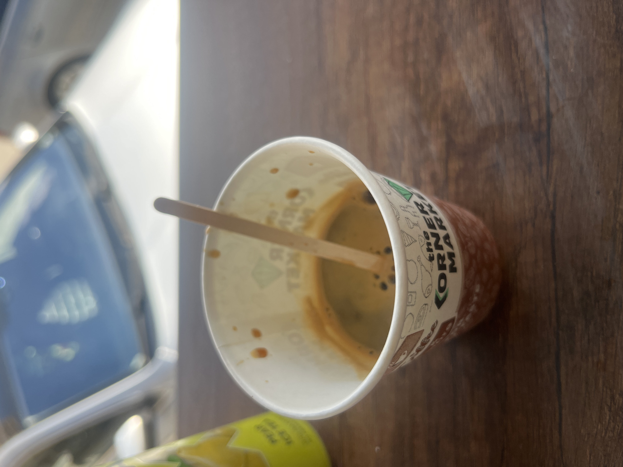 The image shows a paper cup containing a small amount of coffee with a wooden stirrer inside. The cup is on a dark brown wooden surface. Part of a yellow can is visible in the lower left corner of the frame. The blurry image of a car window is visible on the left. The inscription on the cup's side contains the words "THE CORNI" and "MA". No people are visible. The location cannot be confirmed based on visual information.