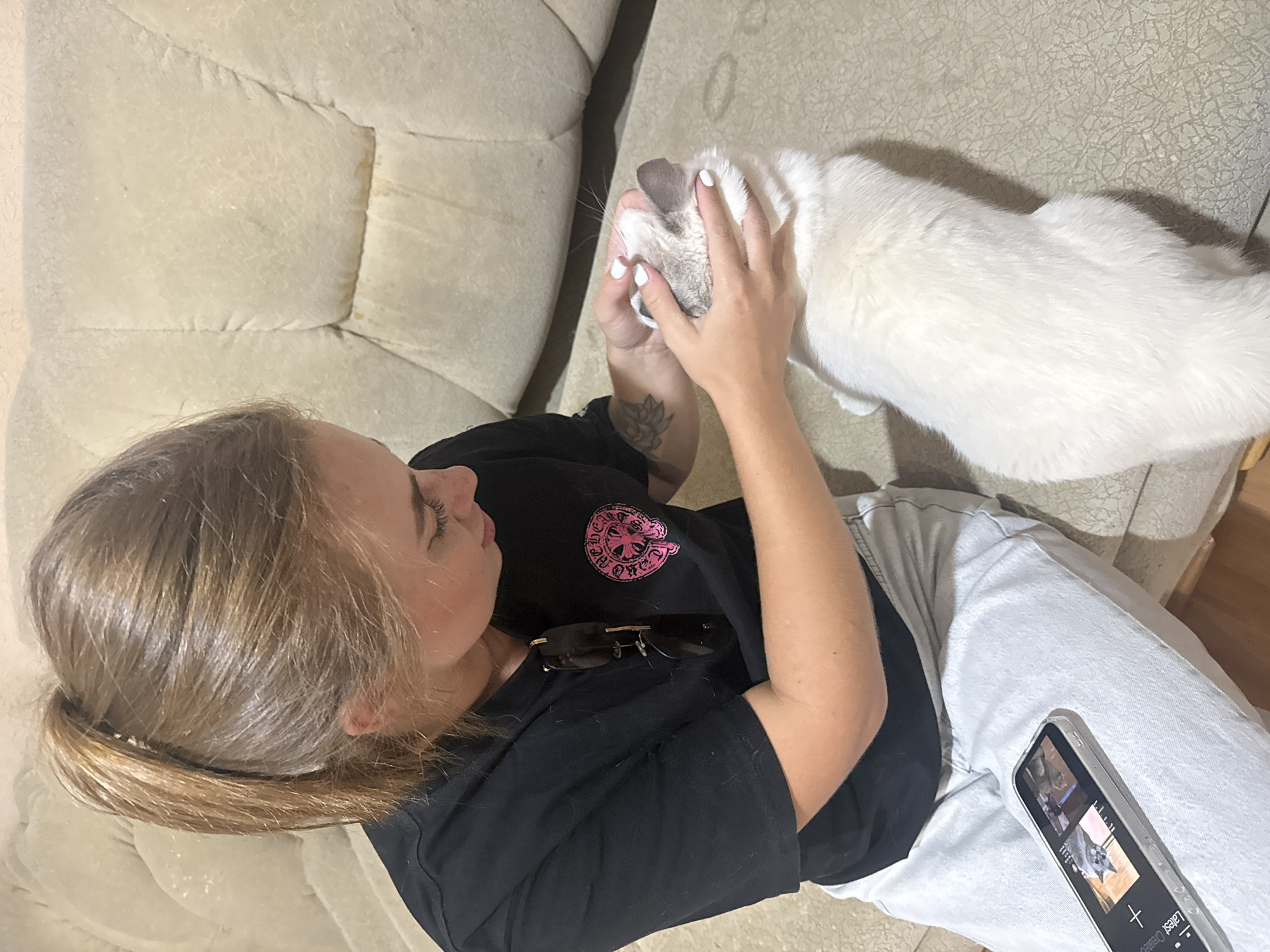 A young woman with a blonde ponytail is sitting on a beige sofa, petting a white cat. She is wearing a black t-shirt and light-colored pants. Sunglasses are clipped to her t-shirt. A smartphone is resting on her lap, displaying images. The background shows a textured beige sofa. The scene is likely indoors, possibly a living room. There is no indication of the city of Dnipro, Ukraine.