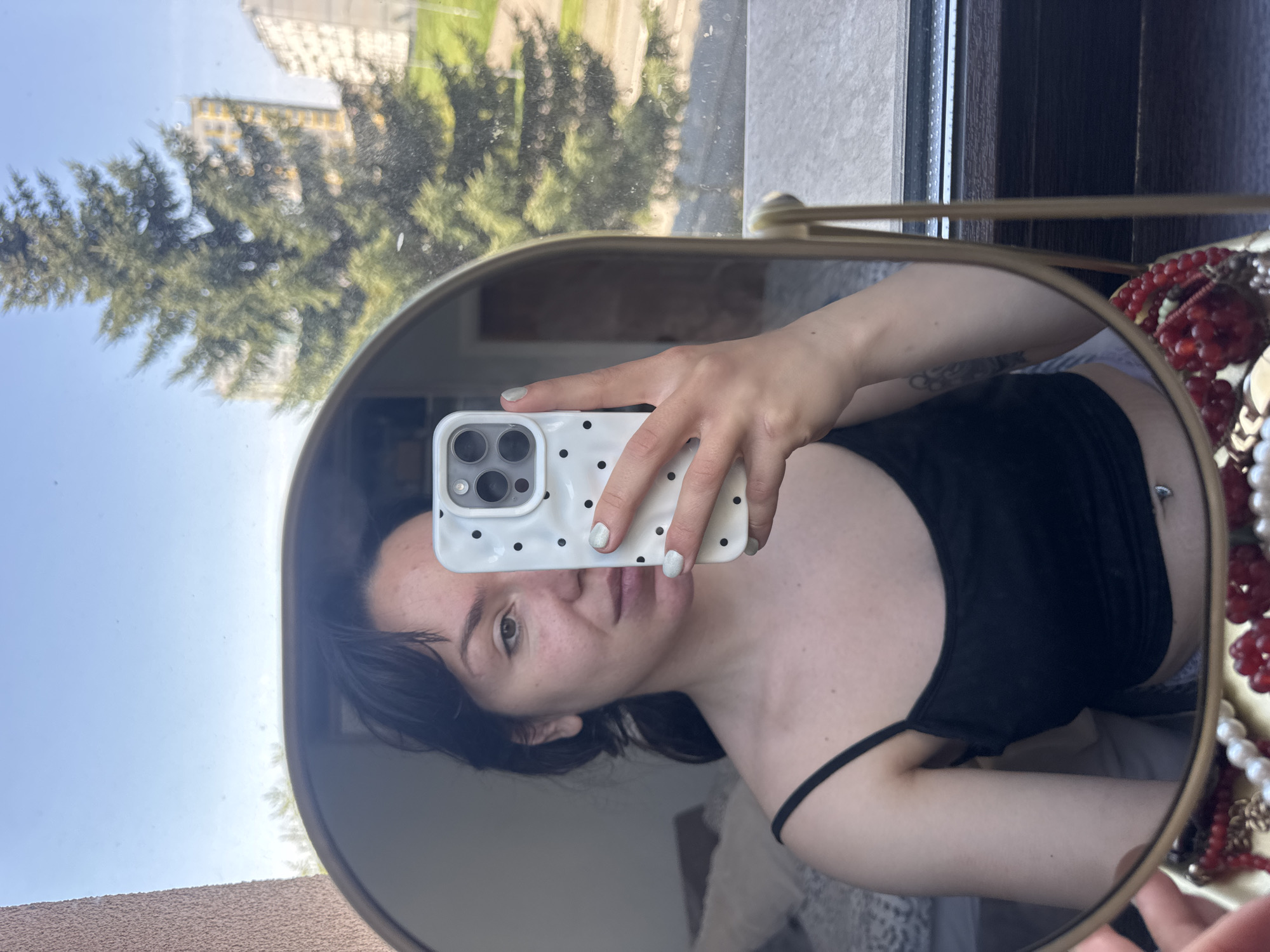 A young woman is captured in a mirror selfie, oriented vertically as if rotated 90 degrees clockwise. She has dark hair and a neutral, calm expression, looking directly at the camera. She wears a black tank top or camisole, and her nails are painted a light, possibly glittery, shade. A navel piercing is visible on her torso, and a subtle tattoo can be seen on her left arm.
She is holding a white smartphone with a black polka-dot case, showcasing its prominent camera array. The selfie is taken in a gold-framed, oval-shaped mirror. Reflected in the mirror, behind the woman, is a clear blue sky, a large green coniferous tree, and parts of light-colored buildings, indicating a bright, sunny daytime setting seen through a window. To the right, outside the mirror's frame, several strands of jewelry are visible, including red beaded necklaces and white/cream pearl-like strands. Elements of the room's interior, such as a textured light-brown surface on the left and dark wooden features on the right, are also partially seen outside the mirror. The setting appears to be an urban or suburban residential area in Burgas, Bulgaria. There is no visible text.
