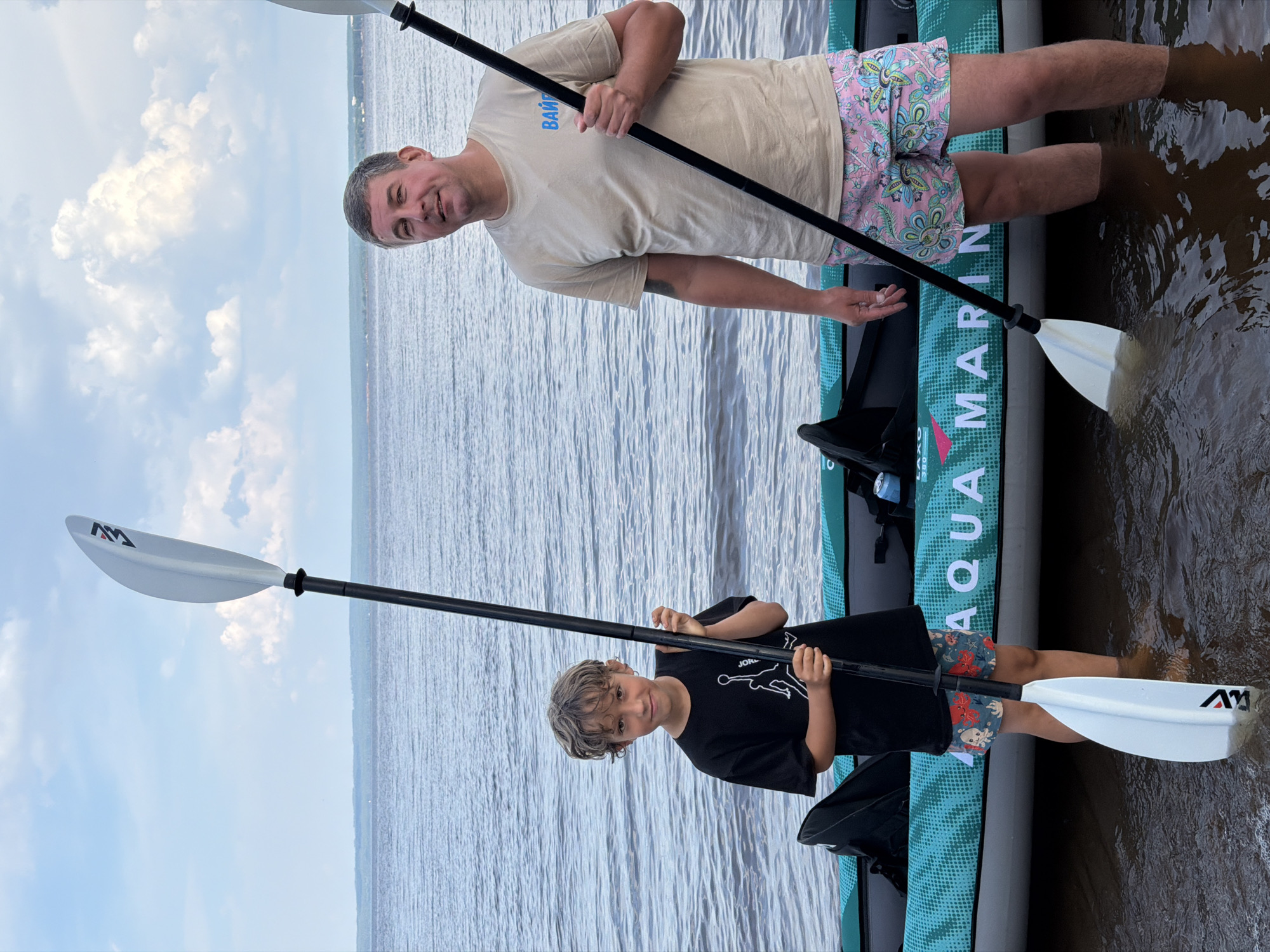 An adult male and a young boy stand in shallow water next to an inflatable kayak on a cloudy day. The boy, on the left, wears a black t-shirt with a white graphic of a jumping figure and floral-patterned shorts. He holds a white paddle with a black shaft, its blade submerged in the water. The man, on the right, wears a light tan t-shirt with blue lettering on the left chest and multi-colored floral-patterned shorts. He holds a black paddle with a white blade that is also submerged. The kayak is teal with white lettering that reads "AQUA MARINA". The background shows a body of water and a hazy horizon line with distant trees and buildings. The location is identified as Noviy, Russia.