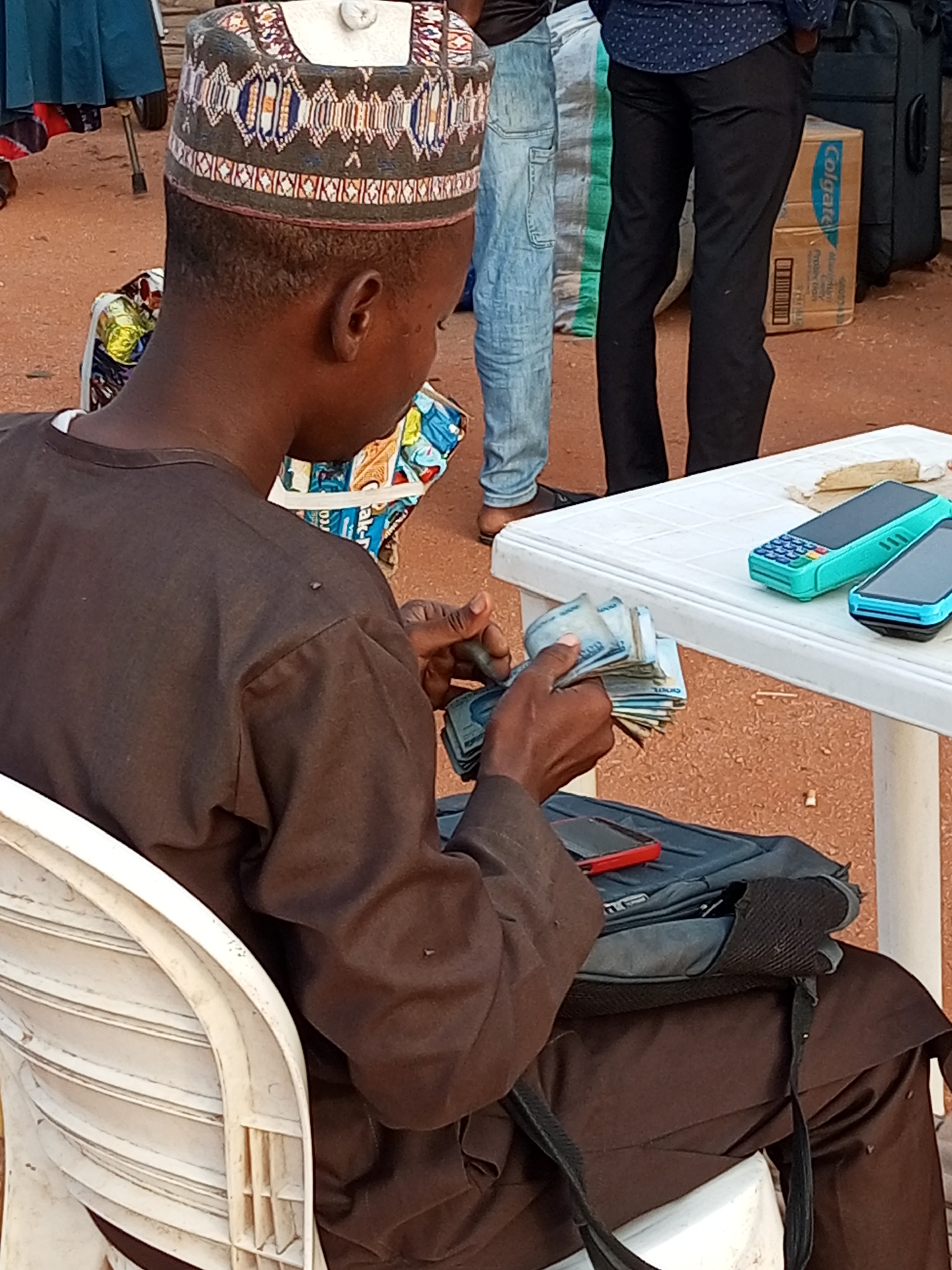 In Gombe, Nigeria, a young Black male, identified as the primary subject, is seated in a white plastic chair. He is wearing a brown traditional outfit and a patterned Kufi cap. In his right hand, he is holding a fan of Nigerian Naira currency, appearing to count or sort the bills. A dark-colored backpack rests behind him, with a red-cased mobile phone on top of it.
In the background, a white plastic table is set up, holding two mobile payment terminals, one turquoise and the other black and blue. Behind the table, a brown cardboard box labeled "Colgate" is visible, along with luggage. Other individuals are present, one wearing a blue patterned shirt and black trousers, and another in light blue jeans. The ground appears to be sandy or dirt. The scene suggests a commercial transaction or informal market activity.