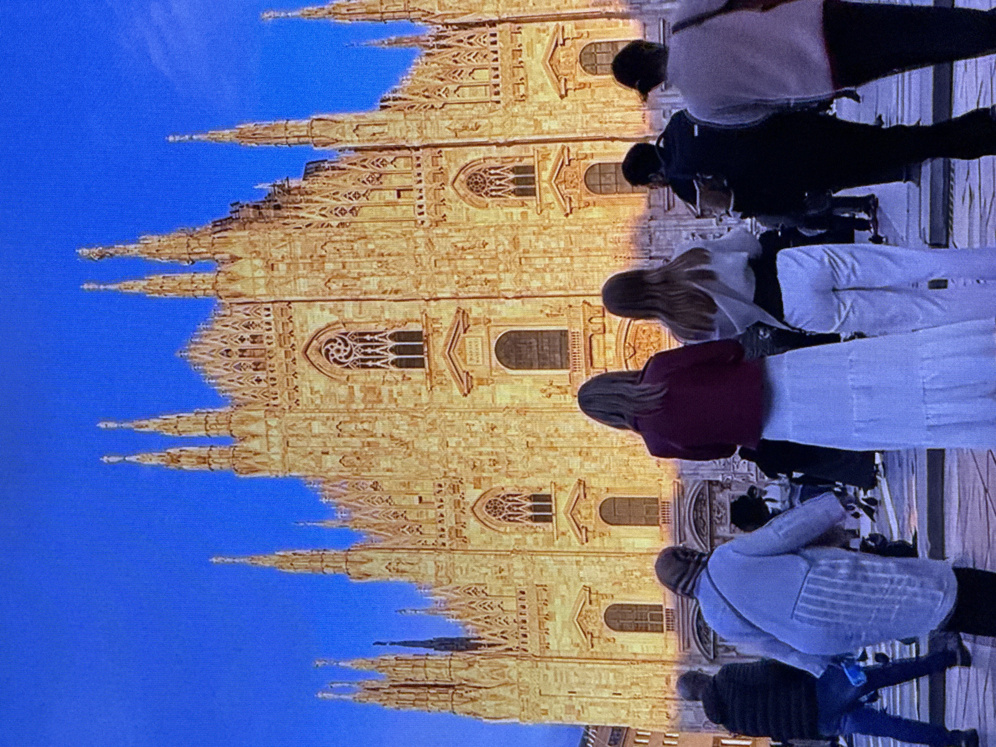 This media file depicts a group of people standing in front of a large, ornate, golden-colored building that appears to be a cathedral or a similar historical structure. The building features intricate architectural details, including pointed spires, arched windows, and decorative carvings, suggesting Gothic or a similar architectural style. The sky is a vibrant, clear blue, indicating daytime and likely fair weather.
Several individuals are visible, appearing to be tourists or visitors. They are dressed in casual clothing, with varying attire suggesting a temperate climate. The people are positioned as if observing or posing for a photograph against the backdrop of the grand building. One person in the foreground wears a light-colored jacket and scarf, another a dark jacket, and a third a dark-colored top. Further back, other figures are less distinct. The composition is vertical, with the building dominating the frame and the people forming a line along its base. The image has a slight tilt, and the bright lighting suggests it's either daytime or the building is illuminated.
Although the prompt mentions Dubai, United Arab Emirates, the architectural style of the building does not strongly align with typical structures found in Dubai. The building's design is more characteristic of European Gothic cathedrals, such as the Duomo di Milano in Italy. There is no visible text in the image.