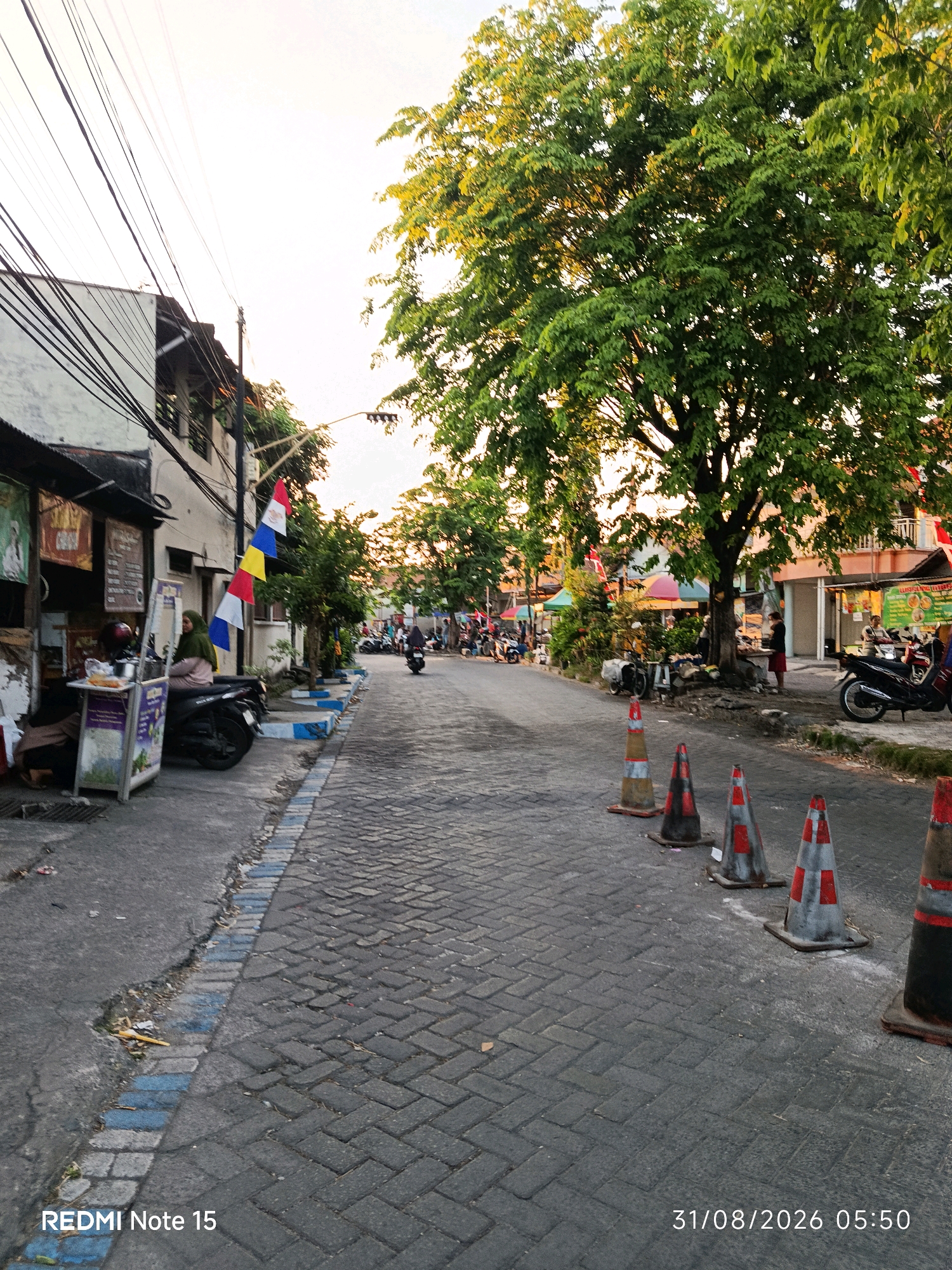 The image depicts a street scene in Keboansikep, Indonesia, at dawn. The street is paved with brick and lined with shops and trees. Several motorcycles are parked on the side of the road, and a woman wearing a hijab is sitting on one of them. Traffic cones are placed along the side of the road, indicating a possible temporary closure or construction. The sky is bright, suggesting the sun is about to rise. The overall impression is that of a quiet, ordinary morning in a residential area.