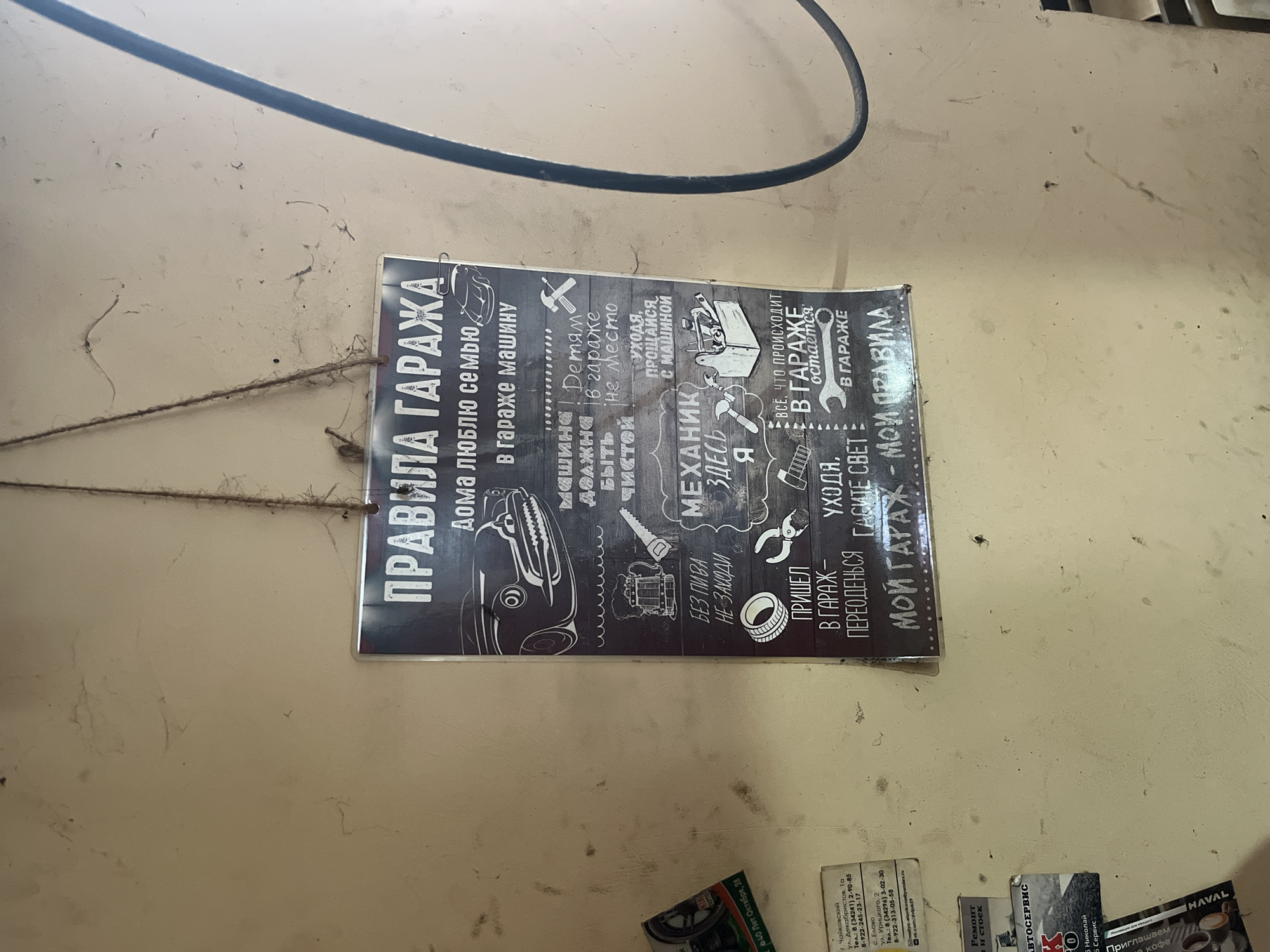 The image captures a dirty, light-colored wall, likely inside a garage or workshop. A dark cable or hose runs horizontally across the upper portion of the wall.
The central subject is a dark-colored sign with white text and illustrations, titled "ПРАВИЛА ГАРАЖА" (Garage Rules). The sign is attached to the wall, possibly with string or twine visible on the left. The rules, presented with a humorous tone, include:
- "At home I love family, in the garage - my car."
- "The car must be clean."
- "Loose parts have no place in the garage."
- "Leaving, say goodbye to the car."
- "Don't come in WITHOUT beer."
- "MECHANIC HERE I am."
- "Everything that happens in the garage stays in the garage."
- "Came to the garage - change clothes."
- "Leaving, turn off the light."
- "MY GARAGE - MY RULES."
At the bottom right, several partially visible business cards or advertisements are affixed to the wall. One prominent card for an "АВТОСЕРВИС" (Auto Service) lists contact information for locations in Chaikovsky city (ул. Декабристов, 1а; Tel: 8 (34241) 2-90-85, 8-922-245-23-17) and Yelovo village (ул. Урицкого, 2; Tel: 8 (34296) 3-02-30, 8-922-313-05-58; email: nastya-elovikova@yandex.ru). Other visible text snippets from these cards mention "Ремонт и стоек" (Repair and struts), "HAVAL", and "Николай Сервис" (Nikolai Service) with "Приглашаем кофе" (We invite to coffee).
The overall setting suggests a practical, well-used garage space in Chaikovsky, Russia, with an emphasis on a proprietor's personal rules and local auto services.