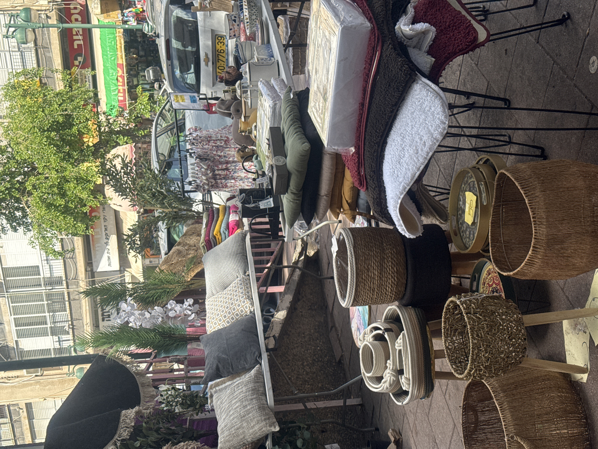 The image shows a street market scene, likely in Ramat Gan, Israel. Merchandise is displayed on tables and racks, including cushions, bedding, towels, and woven baskets. In the background, buildings are visible, interspersed with trees and streetlights. A white van with a license plate number of 40:776-39 is partially visible. A person is standing near the van.
