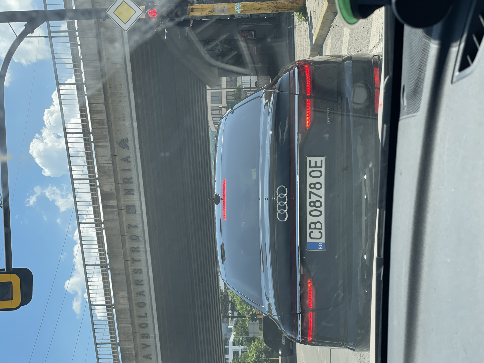 The image shows the rear of a black Audi A8 L Quattro sedan stopped at a traffic light in Sofia, Bulgaria. The car's license plate is visible and reads "CB 0878 OE". Above the car, a concrete overpass with metal railings is visible, with the inscription "AVBOLGARSTROY NRIA" embossed on its side. To the right of the car, a traffic light is visible, currently showing a red signal, and a yellow sign with a diamond shape. In the background, buildings and trees can be seen, suggesting an urban environment. The sky is blue with scattered white clouds. The scene appears to be captured from another vehicle, as the dashboard of a car is visible in the foreground.