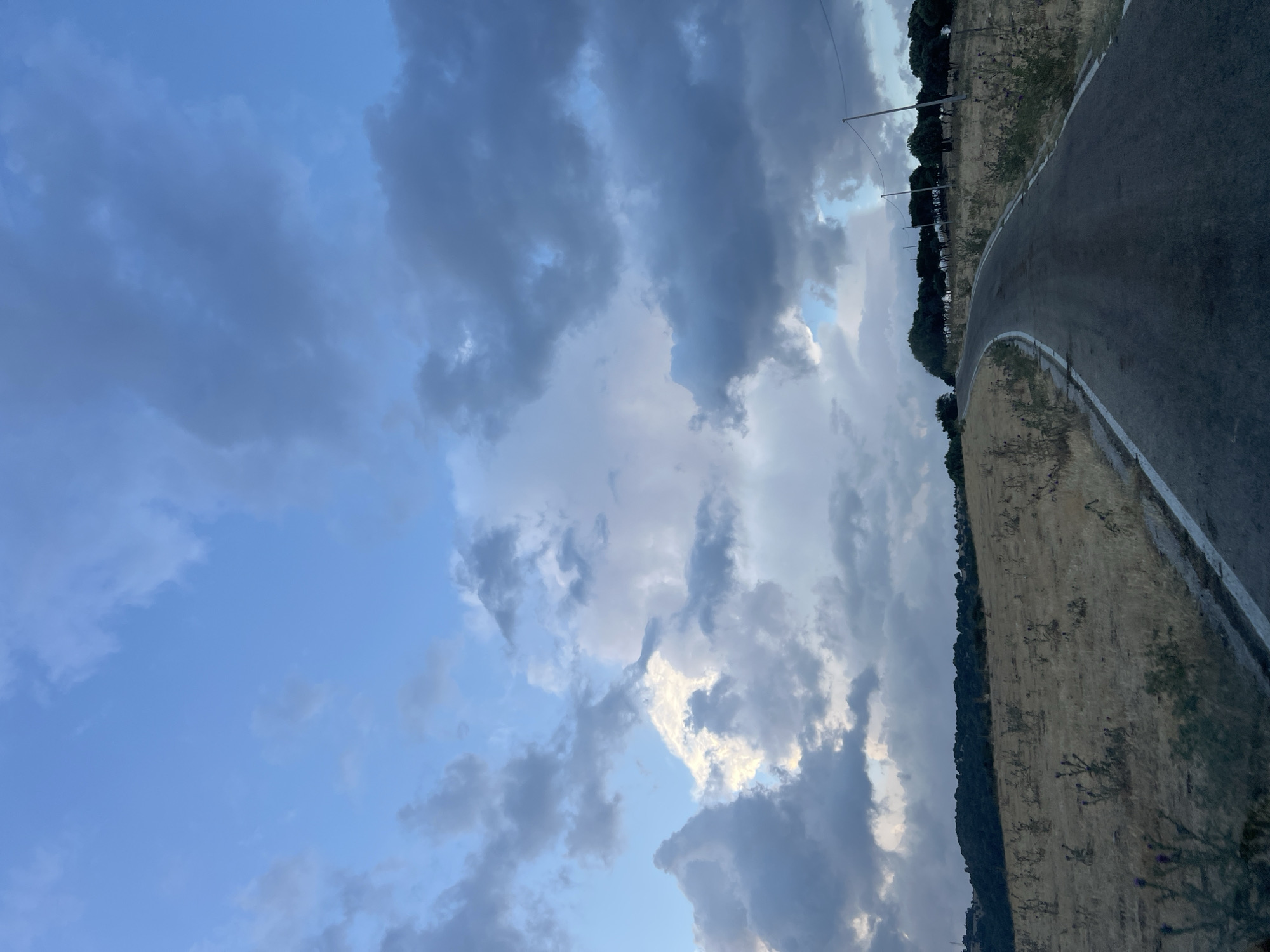 The image depicts a rural road winding through a landscape under a dramatic, cloudy sky. The road is paved and has white lines marking its edges. The surrounding terrain is dry and grassy, with scattered thorny bushes. A line of trees is visible on the crest of a hill in the distance, and utility poles with wires run along the side of the road. The sky is filled with a mix of light and dark clouds, suggesting a dynamic weather condition, possibly at dusk or dawn, with shafts of light breaking through. The scene appears to be in a natural, undeveloped area. There are no people or animals visible in the image. The location in Mattinata, Italy, is not explicitly shown through any recognizable landmarks or signs.