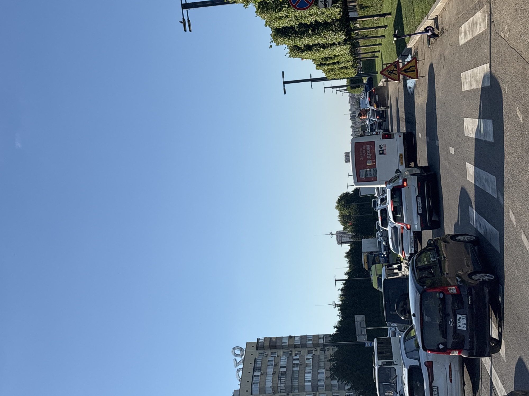The image shows a sunny day in Krasnodar, Russia, with traffic backed up on a multi-lane road. The perspective is from a low angle, looking up towards a clear blue sky and a line of trees and buildings. On the right side of the road, a crosswalk is visible. Several vehicles are stopped in traffic, including cars, a bus, and a truck. Some of the vehicles have visible license plates, and text is present on the bus, reading "За авто иди на дром" and "45". There are also traffic signs visible, including a sign with two figures and a warning triangle indicating road work. A person can be seen standing on the sidewalk next to the road, and a parked electric scooter is also visible. The overall scene suggests a typical urban environment with a traffic jam. The buildings in the background appear to be multi-story residential or office buildings. The sunlight indicates it is daytime, likely morning or afternoon given the clear sky and shadows.