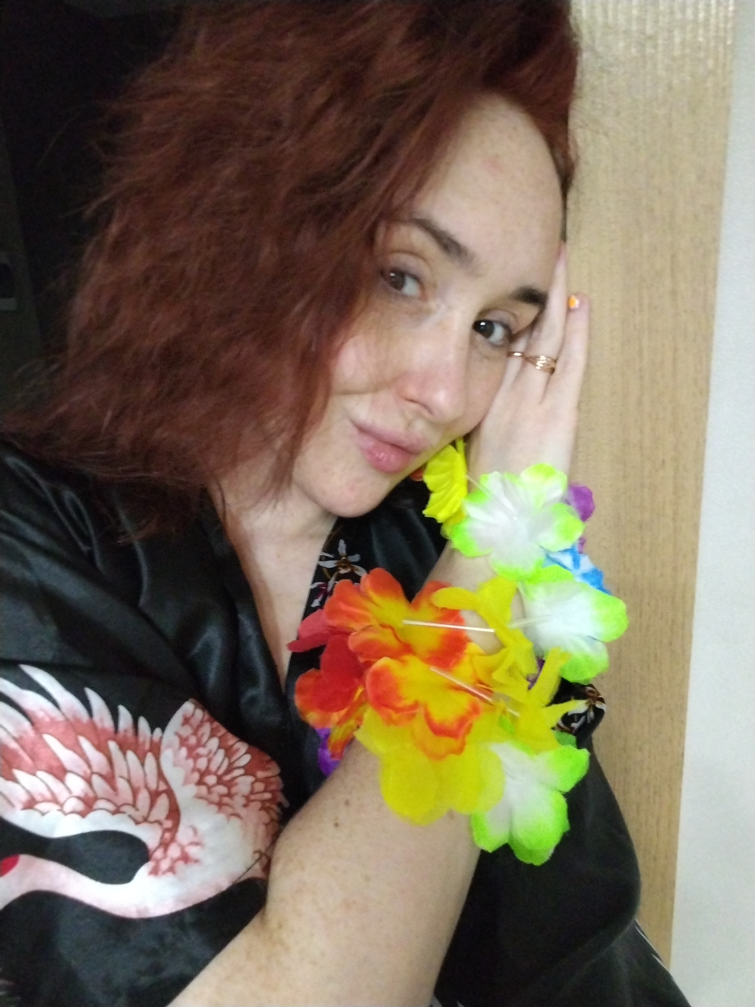 The image presents a close-up portrait of an adult female situated indoors. She possesses fair skin with visible freckles and reddish-brown, wavy hair. Her eyes are directed towards the camera, and a subtle smile is present on her lips. Her left hand, adorned with a gold-colored ring on the ring finger and an orange-painted index fingernail, rests against her left temple. She wears a dark, possibly black, garment featuring a white and reddish-pink bird design, resembling a swan or flamingo, on the upper left chest area. Her right forearm is encircled by several artificial floral leis, displaying a variety of colors including yellow, red, orange, green, blue, and purple. The background immediately to the subject's right is a vertical light-colored surface, appearing to be wood-grained paneling or a door. The background to her left is dark and indistinct. The specific location within the city of Izhevsk, Russia, cannot be determined from the visual information provided in this image.