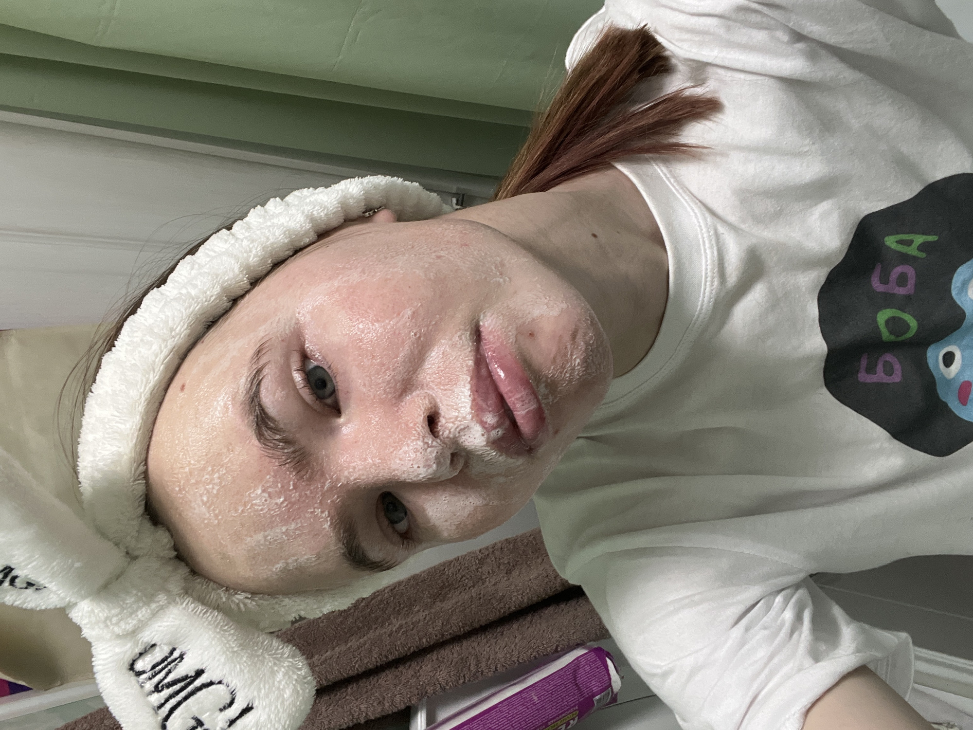 A person with fair skin is shown in a selfie-style image, positioned at an angle to the camera. The individual has a white fluffy headband, featuring the word "OMG!" embroidered on the side. The person's face is covered in a white substance, likely a facial mask. The lips are pink and slightly parted. The background includes a brown towel, a purple package, and a green wall. The subject is wearing a white t-shirt with a dark-colored graphic design on the chest that includes the word "BOBA". The location is Chaikovsky, Russia.