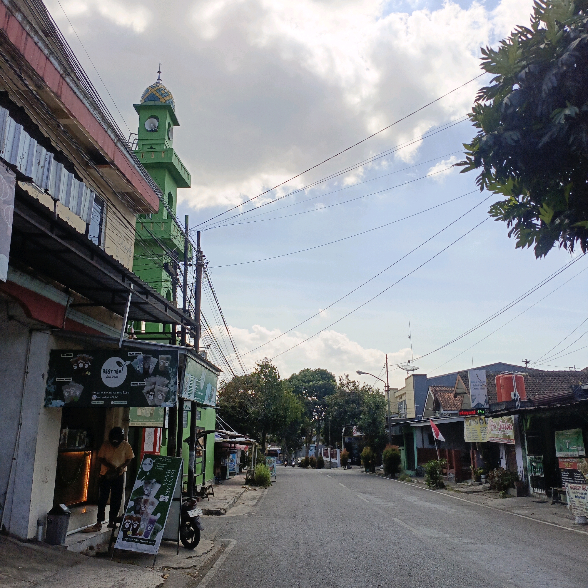 The image shows a street scene in the city of Salatiga, Indonesia. On the left side of the street, there is a green mosque with a dome. Next to the mosque is a shop with a sign that says "BEST TEA". A person is standing inside the shop. There are several motorcycles parked on the side of the street. The street is paved and there are trees on both sides. The sky is cloudy with some blue sky visible. In the background, there are more buildings and trees. The overall scene appears to be a typical street in a small Indonesian city.