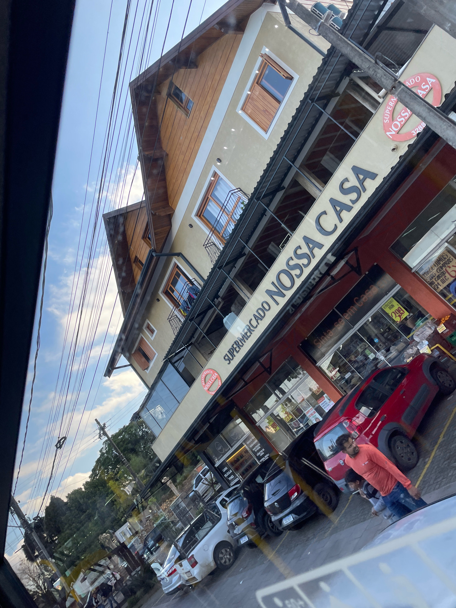 The image shows the "Supermercado Nossa Casa" in Gramado, Brazil, on a clear day. The supermarket is a multi-story building with a light yellow facade and wooden accents on the upper levels. It features large glass windows and balconies. Several cars are parked outside, including a red car and a white pickup truck. A few people are visible, including one man in an orange shirt standing near the red car. Power lines crisscross the sky above the building. The overall scene suggests a commercial area in the city.