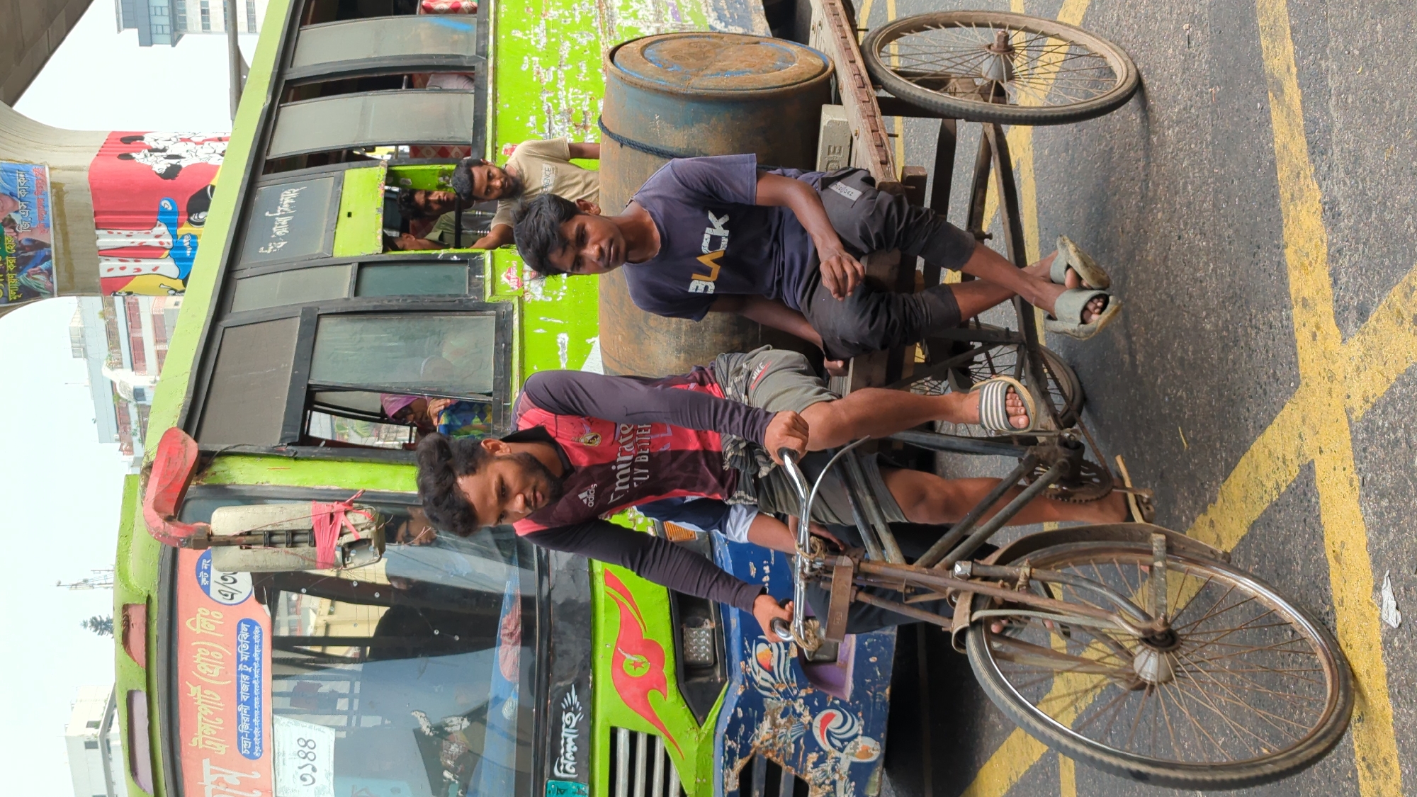 This image depicts a bustling street scene in Dhaka, Bangladesh, captured during daylight hours. The primary subjects are several men and vehicles, set against an urban backdrop.
In the foreground, two men are positioned on and around a sturdy, utilitarian cycle rickshaw or cargo bicycle. One man, wearing a dark red and black "adidas Emirates FLY BETTER" jersey, is at the handlebars, looking downwards. Another young man, wearing a black t-shirt with "BLACK" printed on it and dark pants, sits on the rickshaw's cargo platform, looking directly at the camera with a neutral expression. A rusty barrel is also on the platform. Both men are wearing sandals.
Behind the rickshaw, a vibrant green bus occupies a significant portion of the frame. It shows signs of wear, typical of public transport, and features numerous windows, some of which are open, revealing passengers inside. A side mirror on the bus is visibly tied with red fabric. The front of the bus has Bengali text identifying it as "M Transport (Pvt.) Ltd." with "Route No. A/3" and a detailed route listing "Chandra-Jirani Bazar to Motijheel" and several other locations including "Sreepur," "Gabtoli," "Farmgate," and "Gulistan." The number "3144" is also displayed, likely its vehicle number. On the bus's side, "জি.এস কাকন" (G.S. Kakon) is visible above the windows.
In the background, a concrete overhead structure, possibly a flyover, looms. Beneath it, a partial view of a red billboard with Bengali text "মূল্যায়ণ করা হোকনা" (Let it be evaluated) can be seen. The ground is paved asphalt marked with yellow lines. The scene conveys a candid moment of everyday life in a lively South Asian city.