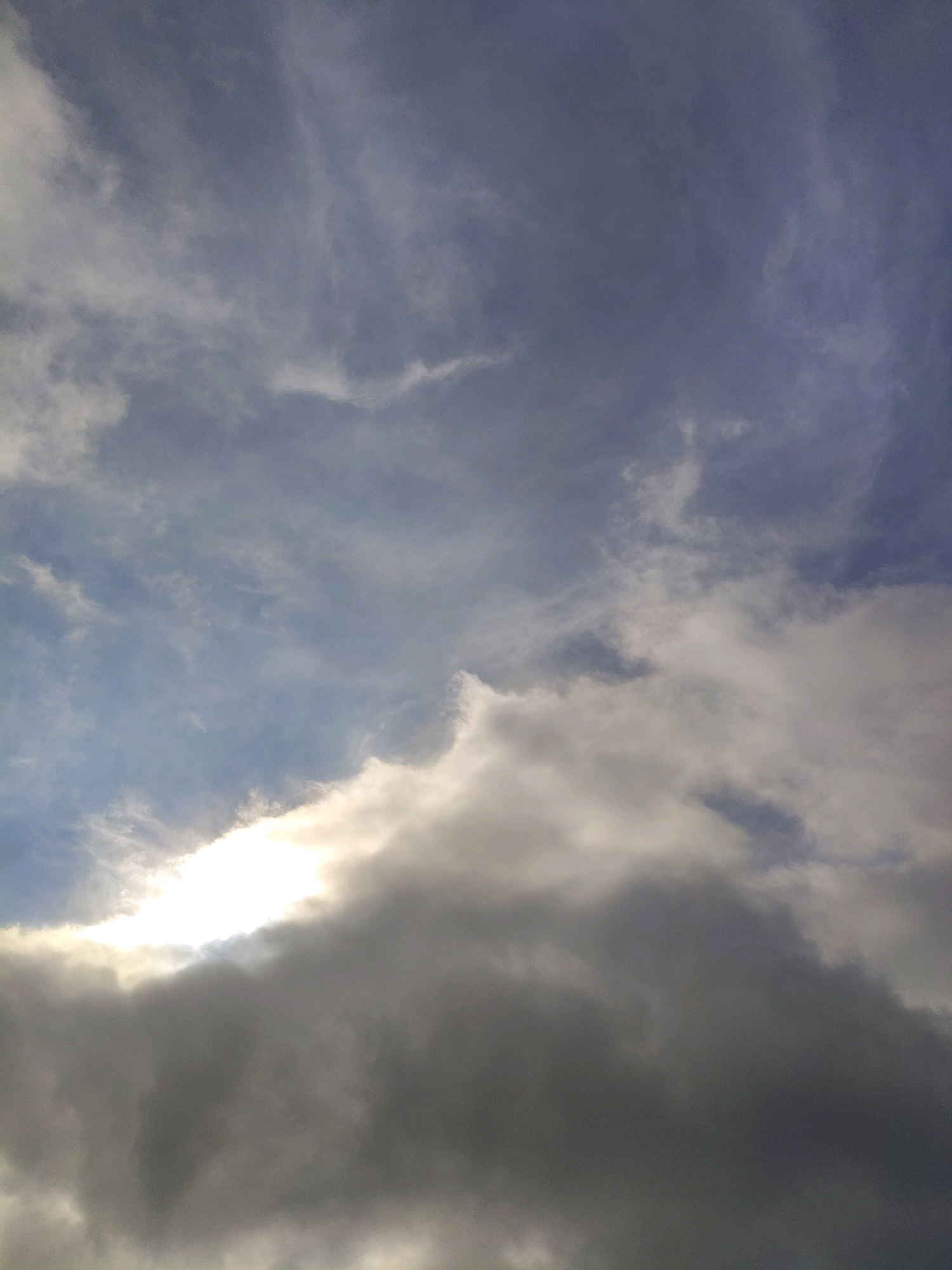 The image shows a sky filled with clouds. A bright light source, likely the sun, is partially obscured by dark, heavy clouds in the lower portion of the frame. Lighter, wispy clouds are visible in the upper part of the image, with patches of blue sky peeking through. The scene is purely atmospheric, with no identifiable city landmarks, people, or objects.