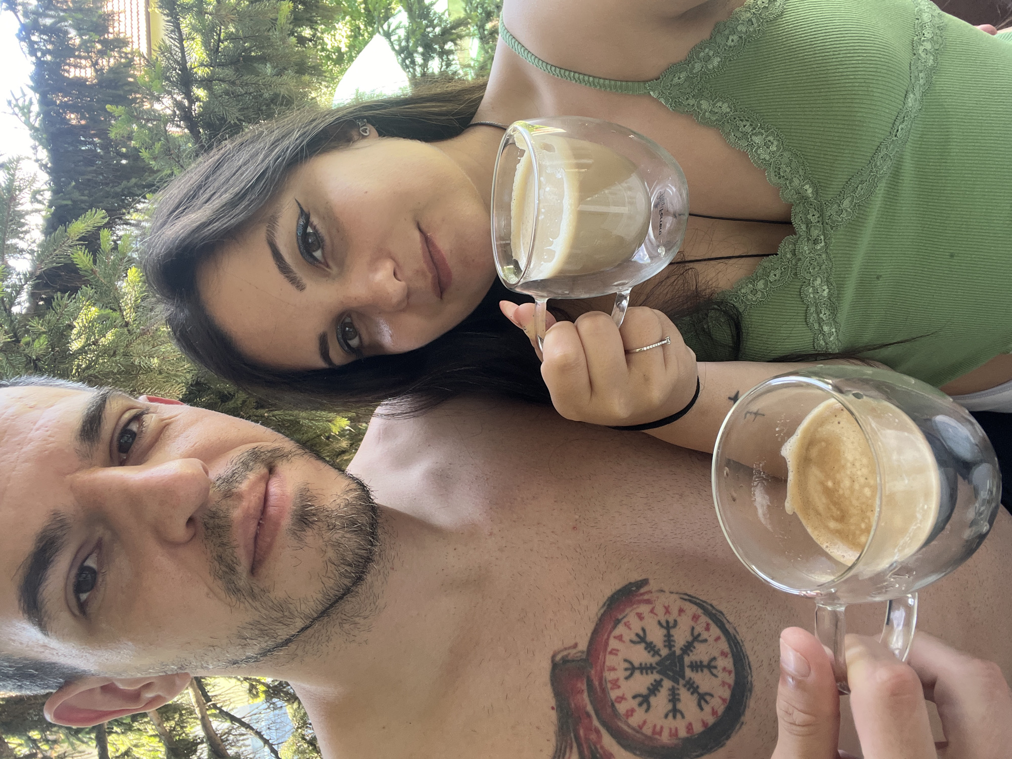 A man and a woman are posing for a selfie, holding coffee cups. The man, with short dark hair and a beard, is shirtless, revealing a tattoo on his chest. The woman, with long dark hair, is wearing a green top and has makeup on. They are outdoors, with trees and a house visible in the background. The scene appears to be from Sofia, Bulgaria, based on context.