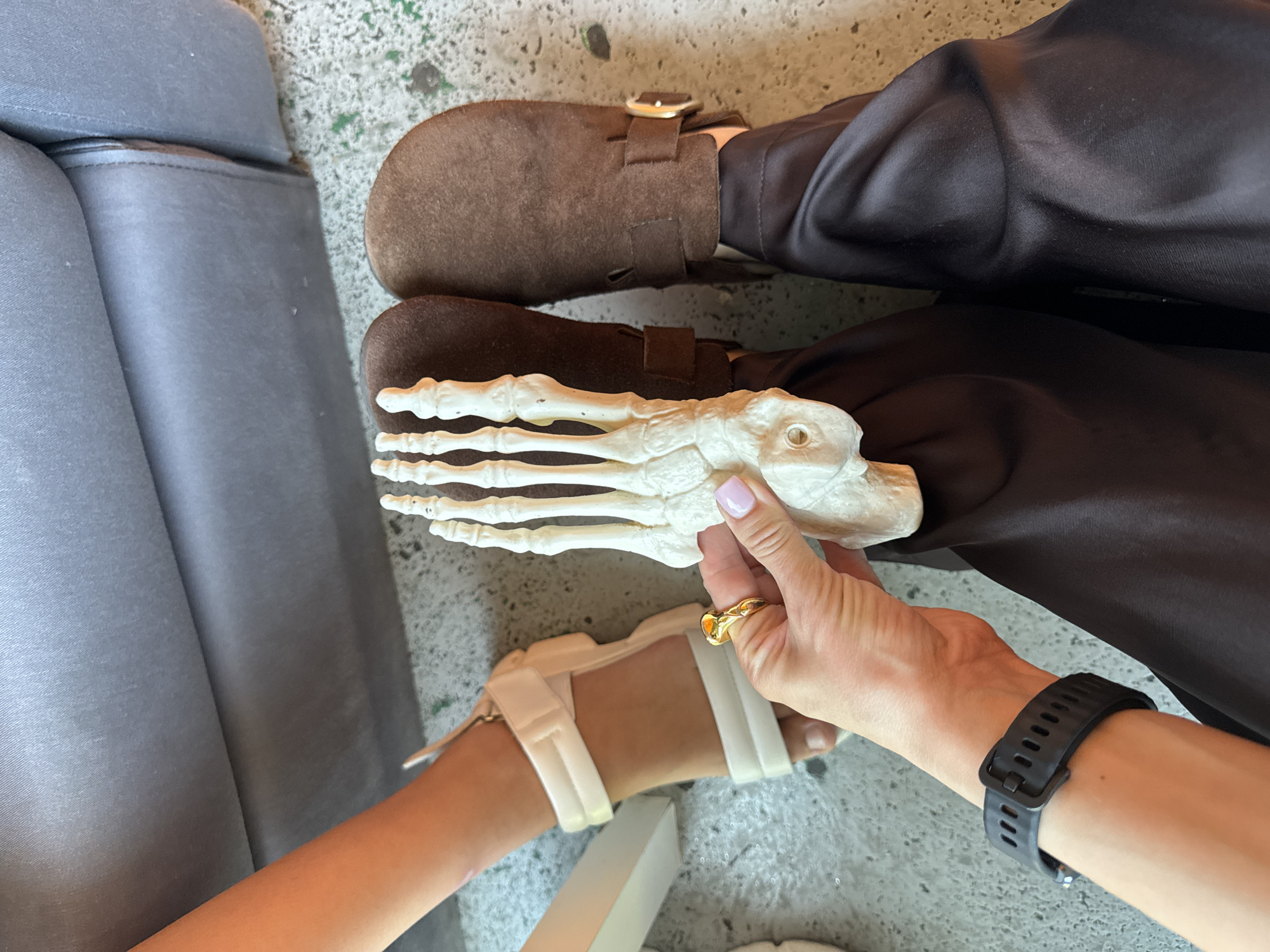 The image shows a close-up, overhead view of two people's legs and feet, with a model of a human foot being held by one of them. One person is wearing white platform sandals and dark satin pants. The other person is wearing brown suede clogs and dark satin pants. The model foot is white and appears to be made of plastic or a similar material. The setting appears to be indoors, with a grey upholstered surface and a speckled floor. There is no indication of the city of Sochi, Russia, or any other specific location in the image. The image is a still photograph and there are no ongoing actions or interactions depicted beyond the placement of the objects.
