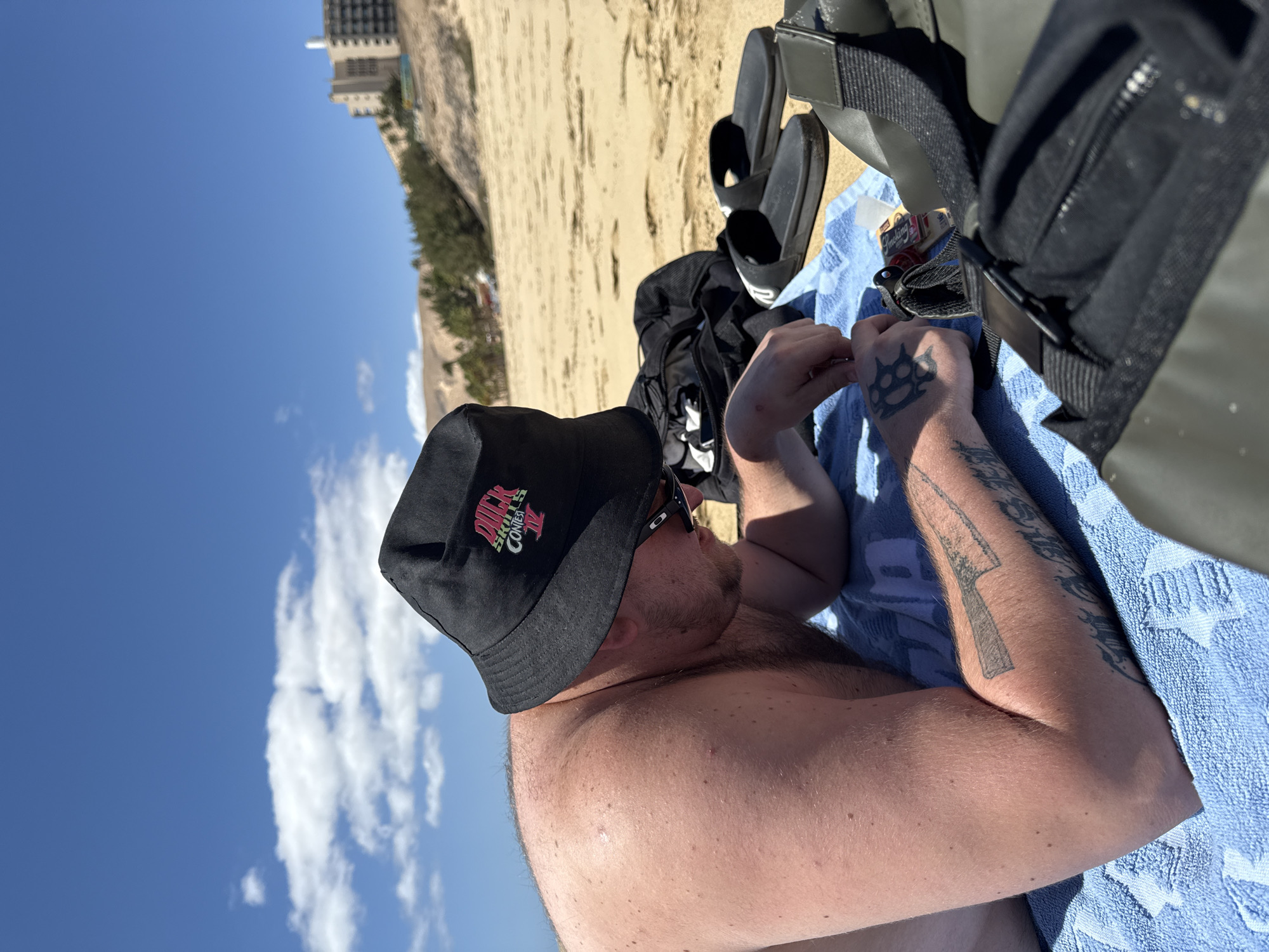 This image, currently displayed on its side, depicts a person relaxing on a sandy beach under a bright, clear blue sky in Pájara, Spain. The central figure is a shirtless man, seen from the chest up, lying on a light blue towel. He is wearing black sunglasses and a black bucket hat. The hat features a pink logo with text that appears to read "DUCK SKATE CONTEST."
His arms are visible, displaying several tattoos, including a knuckle duster design on his left hand and a stylized knife or dagger on his left forearm. Around him, various personal items are scattered on the towel and sand: a pair of black slides, a black duffel bag or backpack, and a pack of "Smoking Deluxe" rolling papers.
The background reveals a wide expanse of light brown sand, leading to distant greenery and a tall building, possibly a hotel or resort, under a sky dotted with a few white clouds. The bright sunshine indicates it is daytime, suggesting good weather for leisure and relaxation on the beach.
