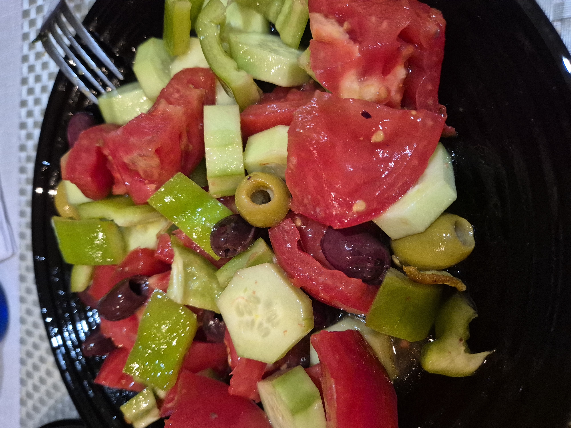 A close-up shot showcases a black plate filled with a colorful Greek salad. Chunks of ripe red tomatoes, bright green bell peppers, and crisp cucumber slices are mixed with Kalamata olives. A silver fork rests on the edge of the plate, ready to be used. The salad is glistening, suggesting it has been dressed, likely with olive oil and a light vinaigrette. The background is blurred, with hints of a checkered pattern and the edge of a blue container, indicating an indoor setting. The lighting is bright, highlighting the fresh ingredients. The scene is forensic and matter-of-fact, with no people or specific location in the city of Unknown, Unknown.